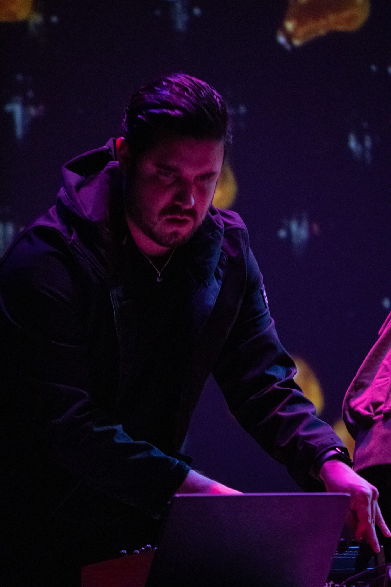 Man with dark hair and beard working on a laptop in a dimly lit environment with colorful lights.