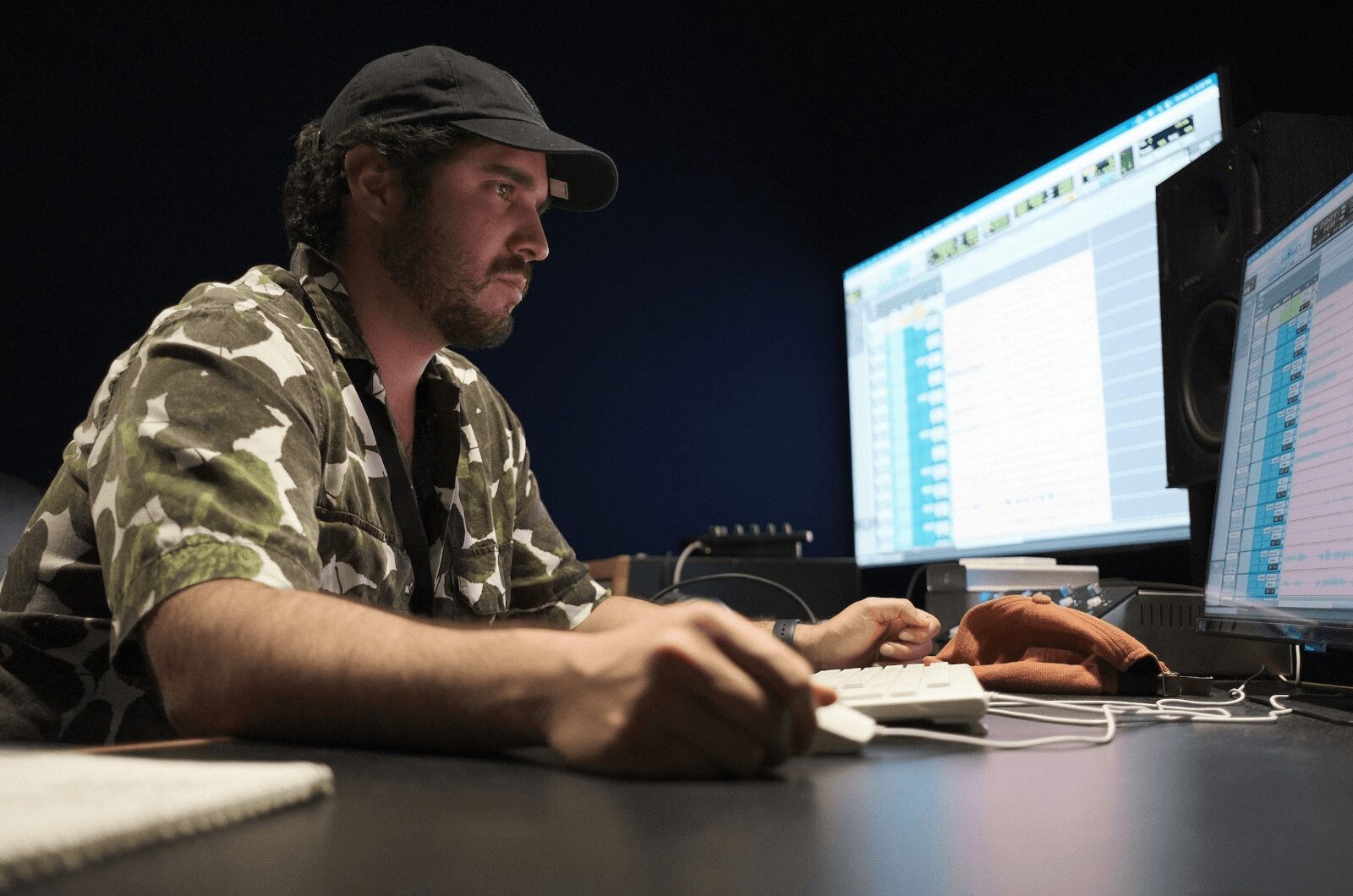 Man with a beard and cap working at a desk with two computer monitors in a dark room.