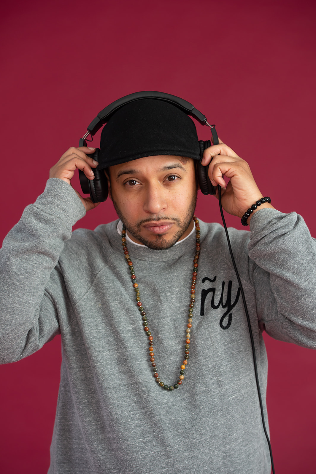 Young man wearing a black cap and headphones, adjusting the headphones with both hands, against a pink background.