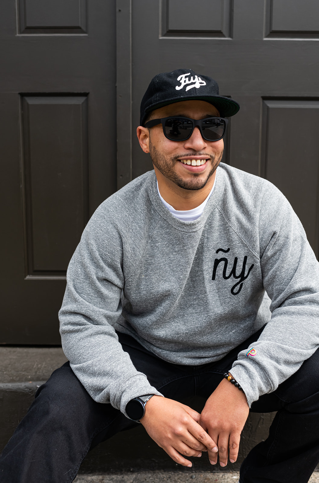 Man sitting outdoors, wearing sunglasses, a black cap, and a gray sweatshirt, smiling at the camera.