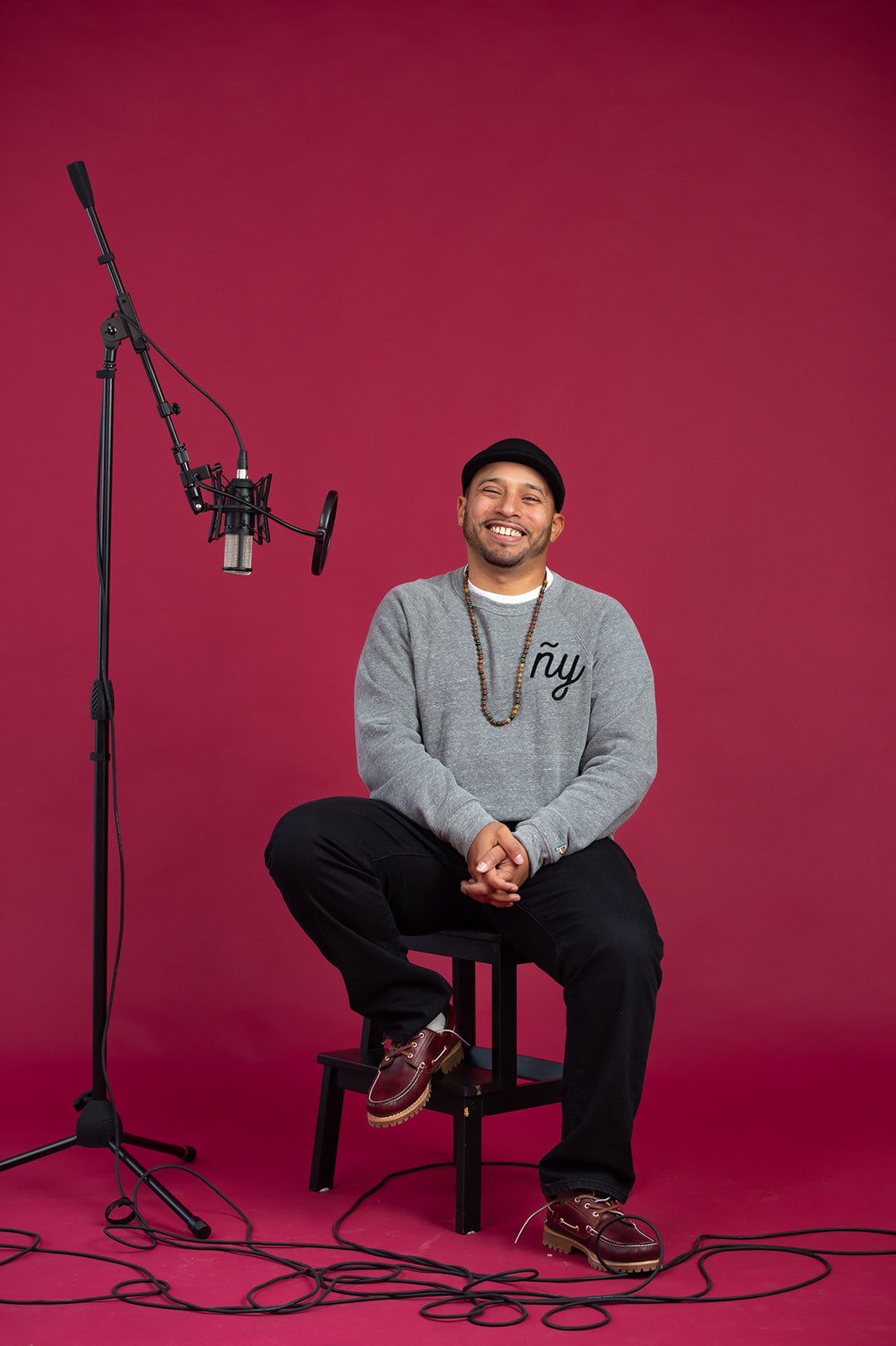 Man sitting on a stool with a microphone stand and cables against a pink background.