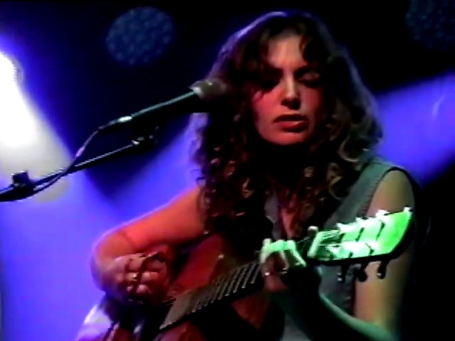 Woman with curly hair playing an acoustic guitar on stage with purple and blue lighting.