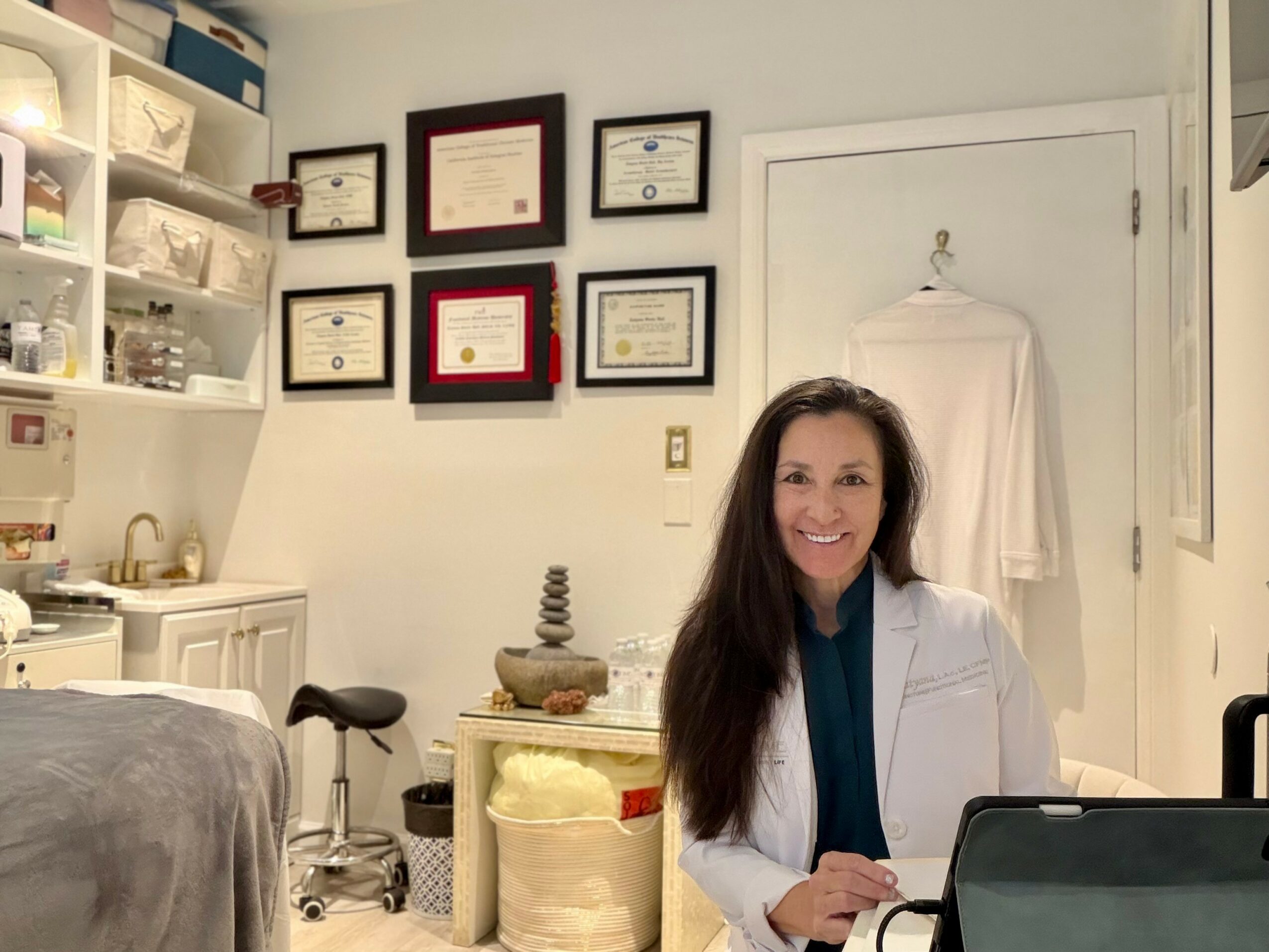 Woman with long dark hair wearing a white coat sitting at a desk with a laptop in a room with framed certificates on the wall.