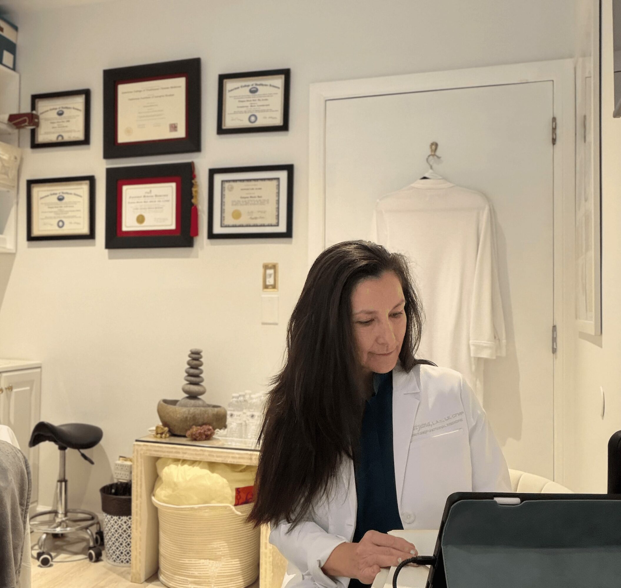 Woman in white coat working at desk in office with framed certificates on wall.