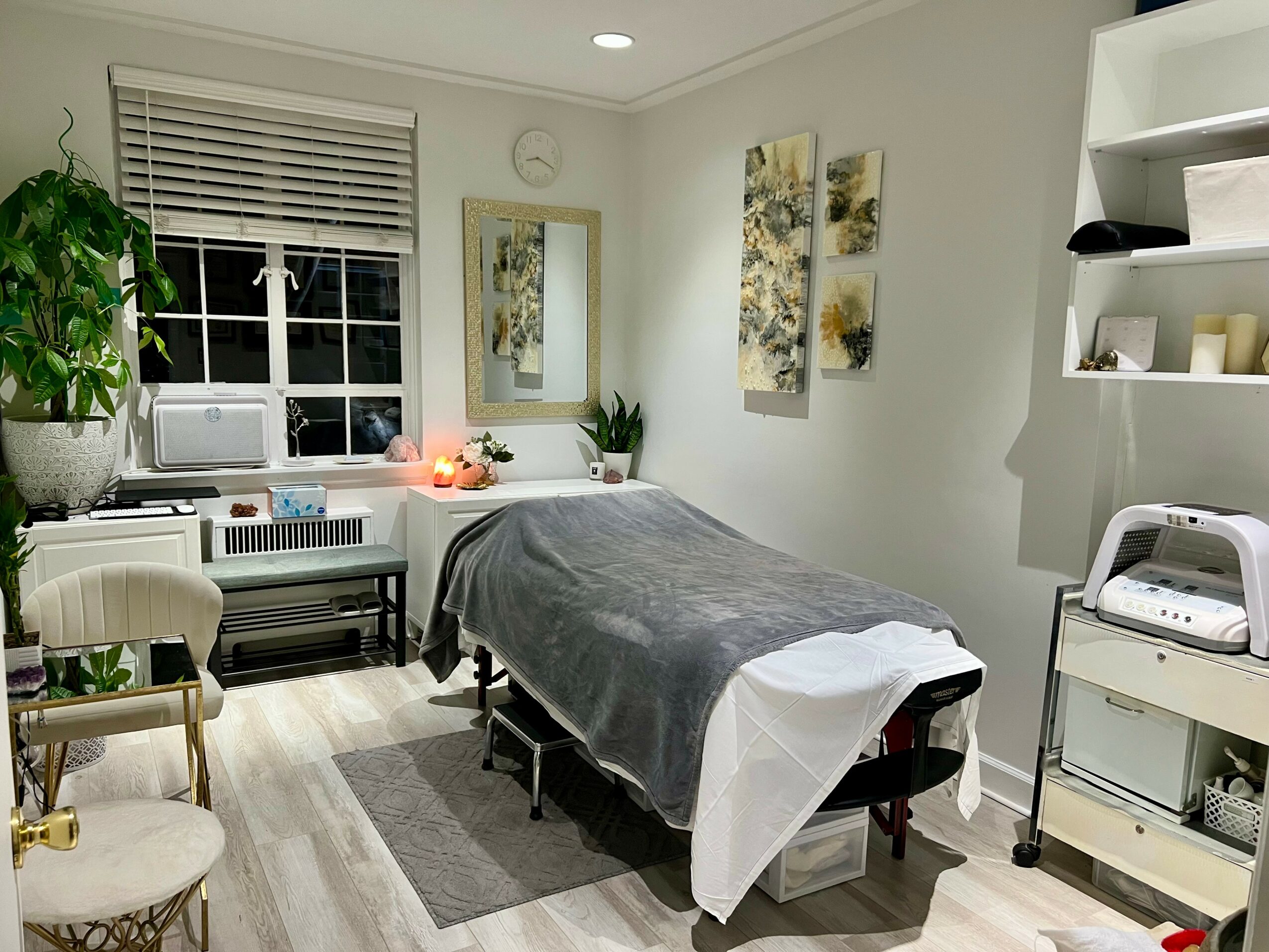 Treatment room with massage table, window, artwork, and shelves with supplies, decorated in neutral tones.