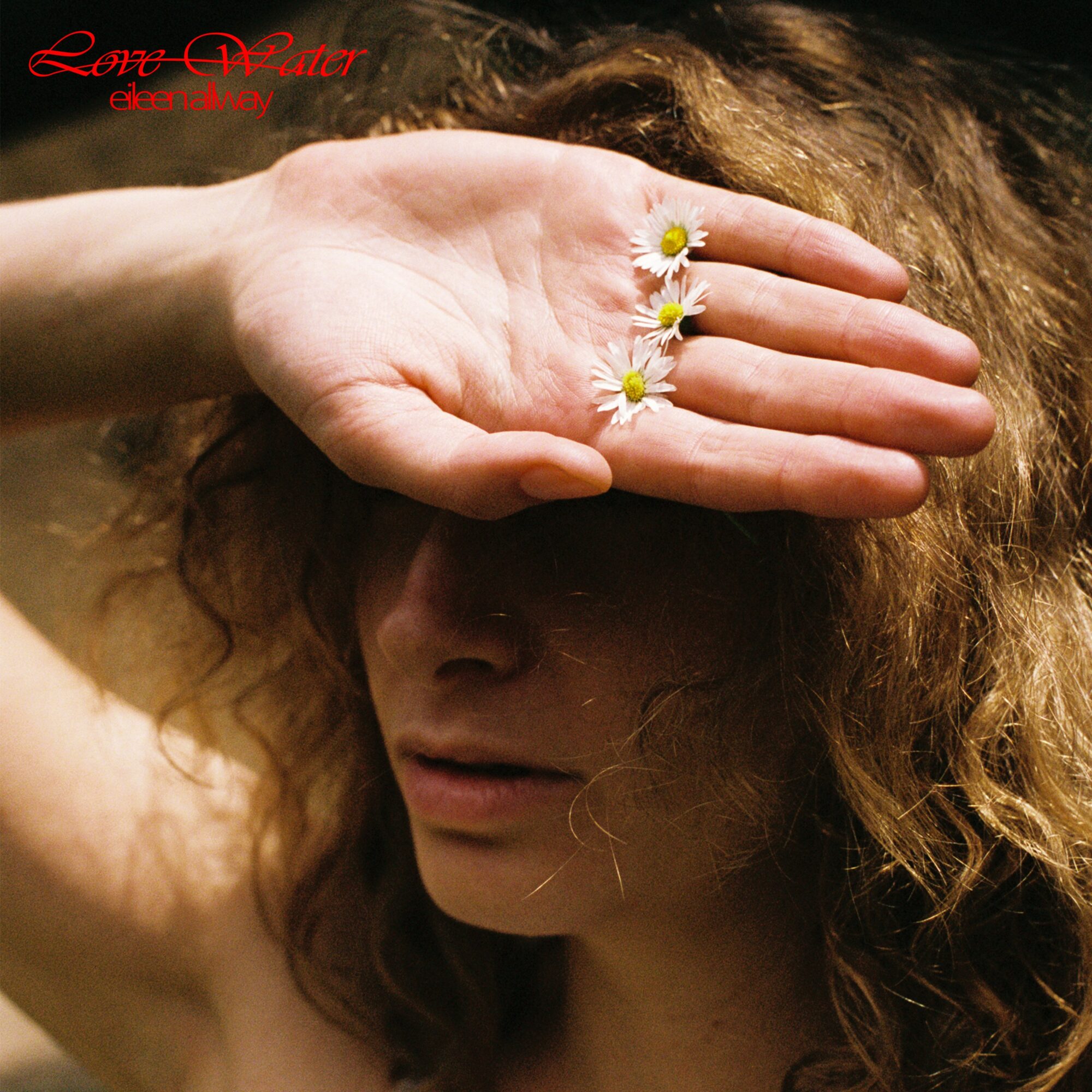 Person with curly hair holds hand with daisies over face, obscuring eyes, in a close-up portrait.