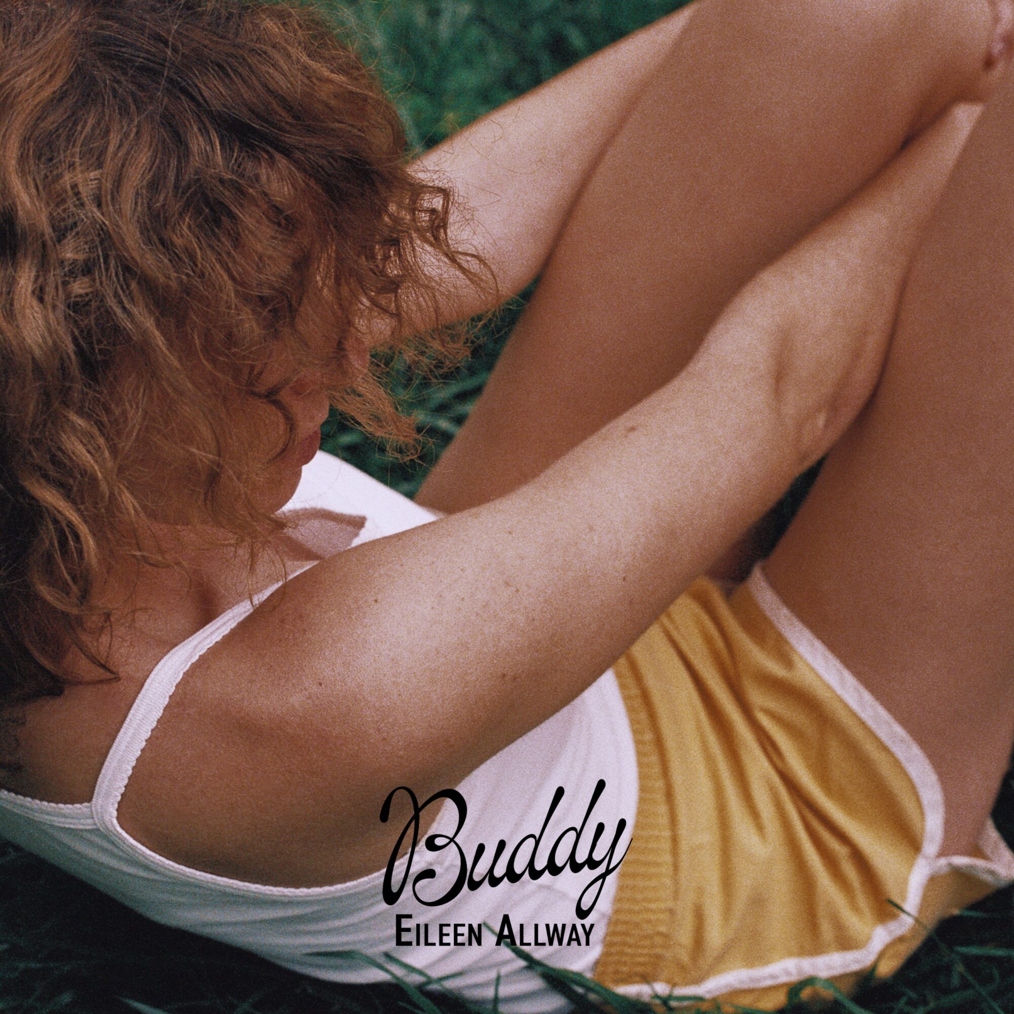 Person with curly hair lying outdoors, wearing a white top and yellow shorts, with arms raised.