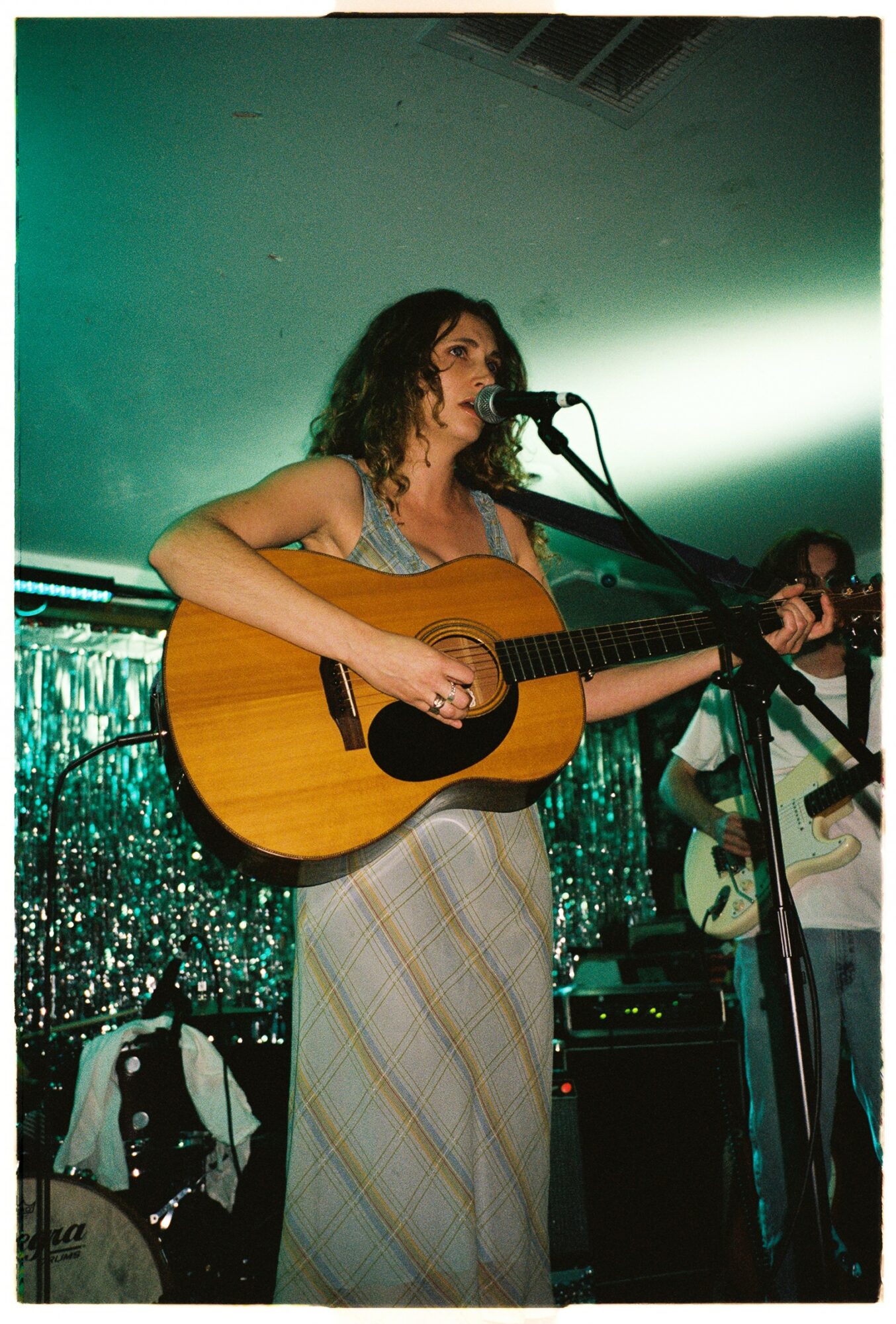 Woman with curly hair singing and playing guitar on stage with a microphone, backdrop of shiny decorations.