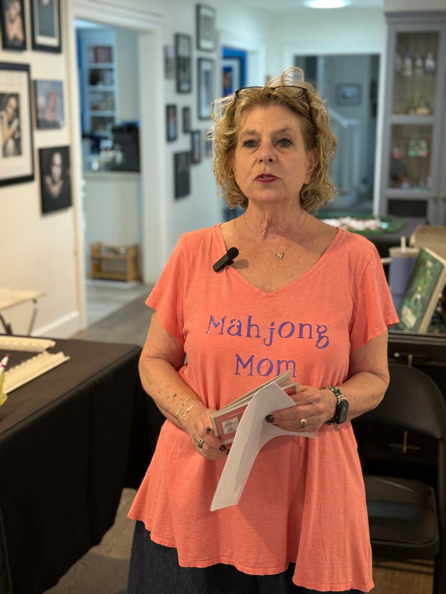 Older woman with curly hair holding papers, standing in a room with bookshelves and framed pictures.