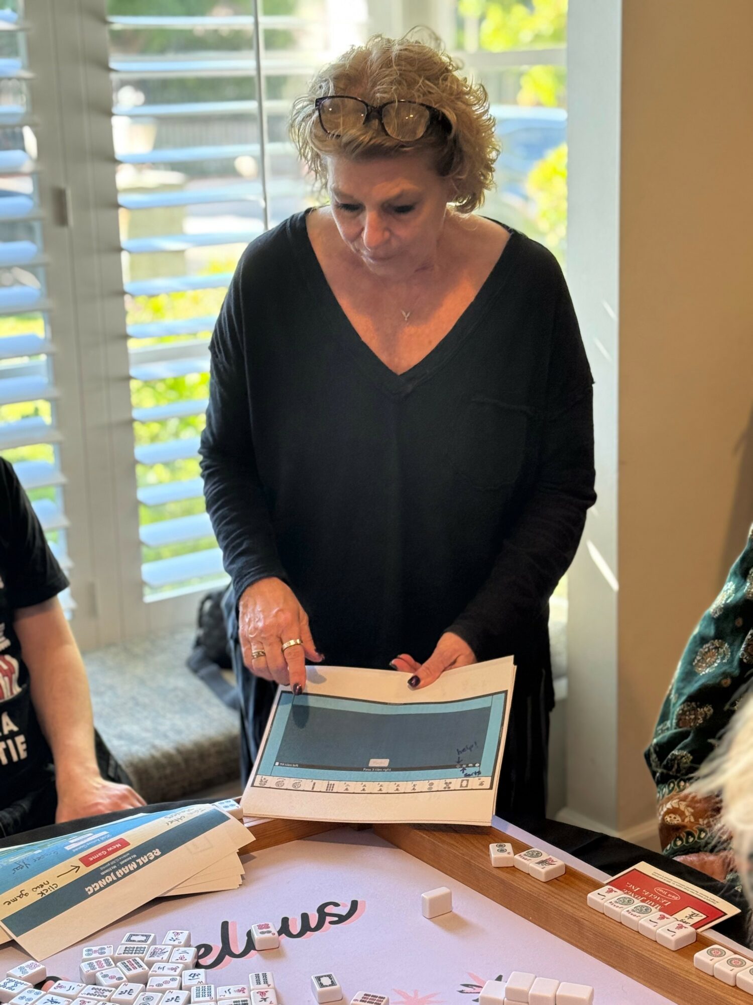 Woman with curly hair and glasses looking at a tablet on a table, with books and papers nearby.