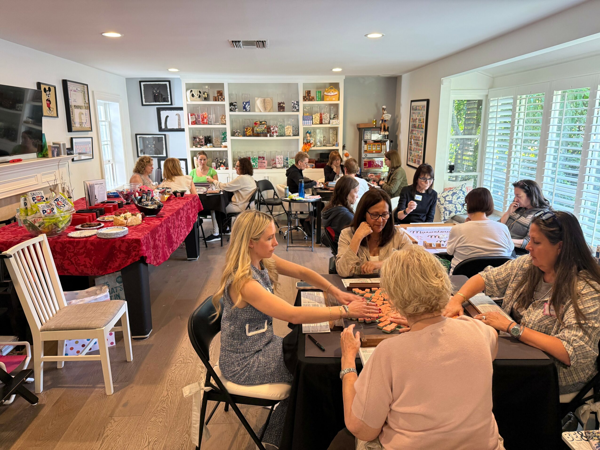 Group of people sitting at tables in a bright room with large windows and bookshelves, engaging in activities.