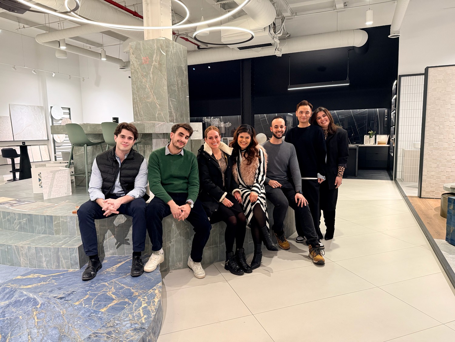 Group of seven people sitting and standing in a modern indoor space with white walls and industrial decor.