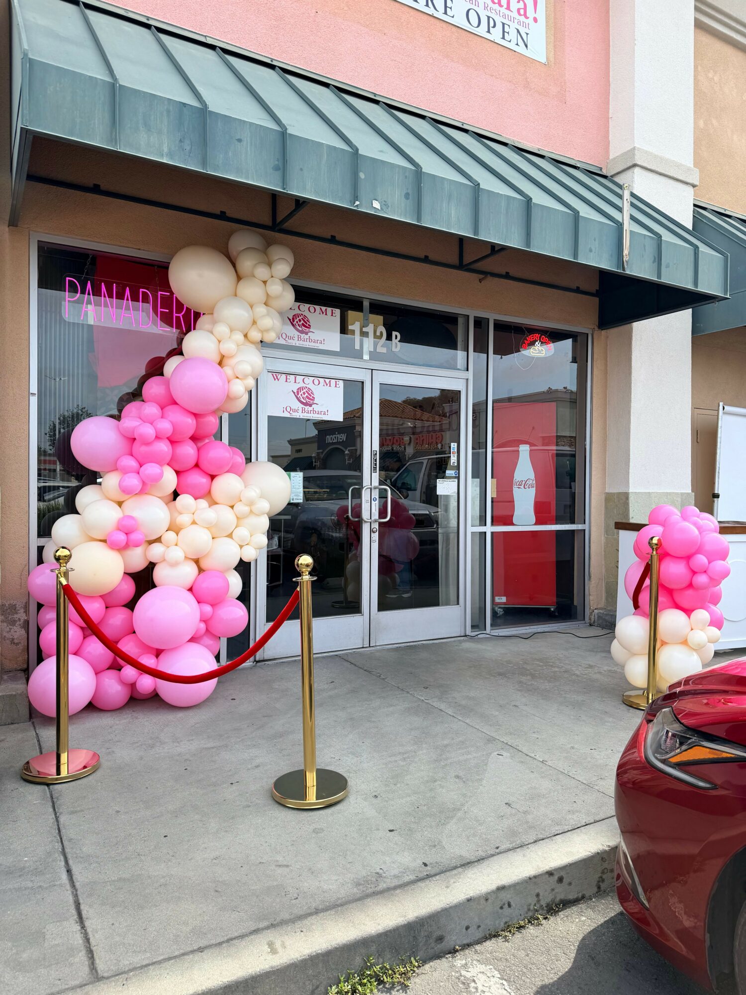 Store entrance decorated with pink, white, and cream balloons, gold stanchions, and red velvet rope, outside shopping mall.