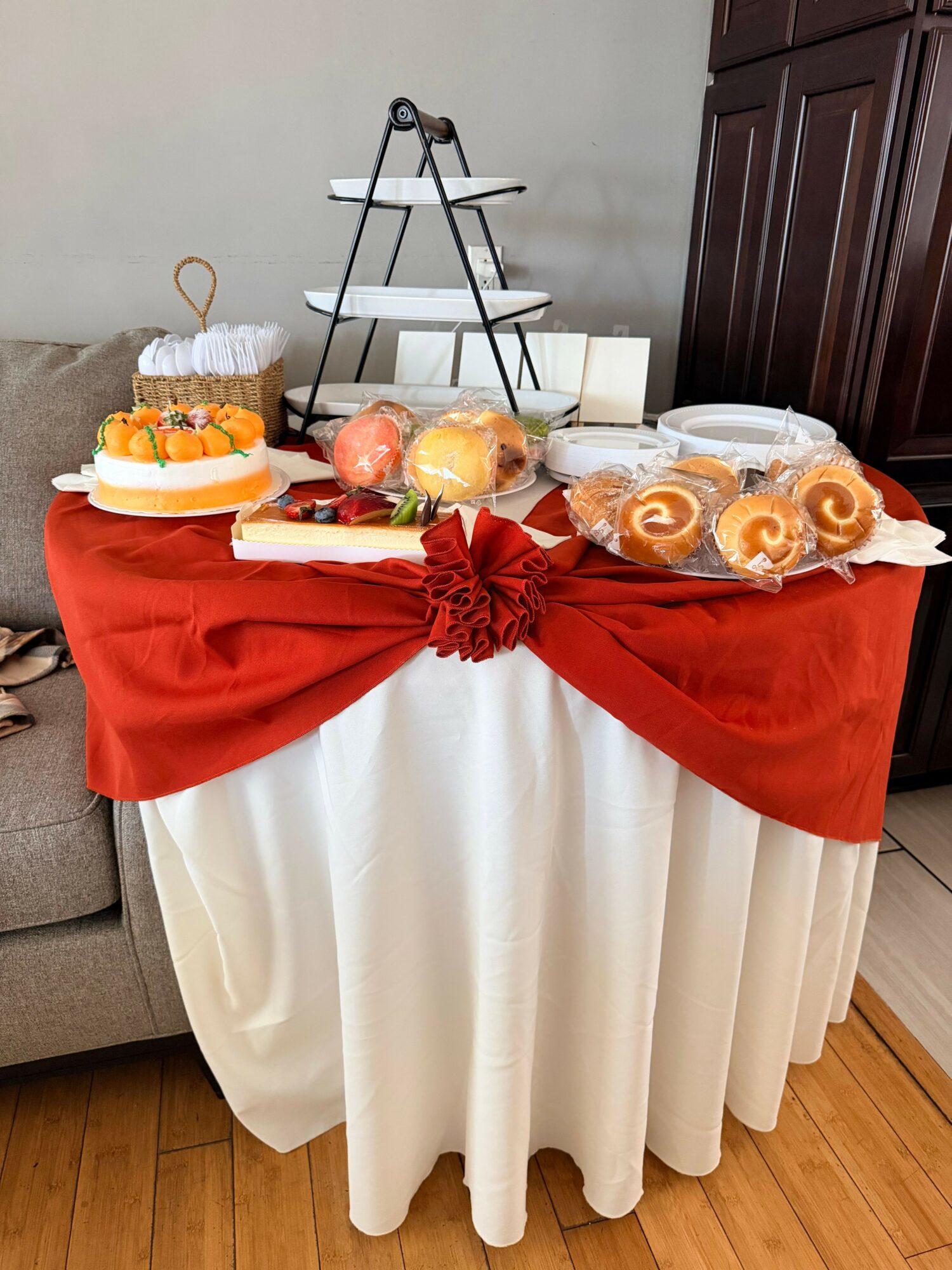 Table with red and white cloth, decorated with baked goods, apples, and a red bow, against a wall and cabinet.