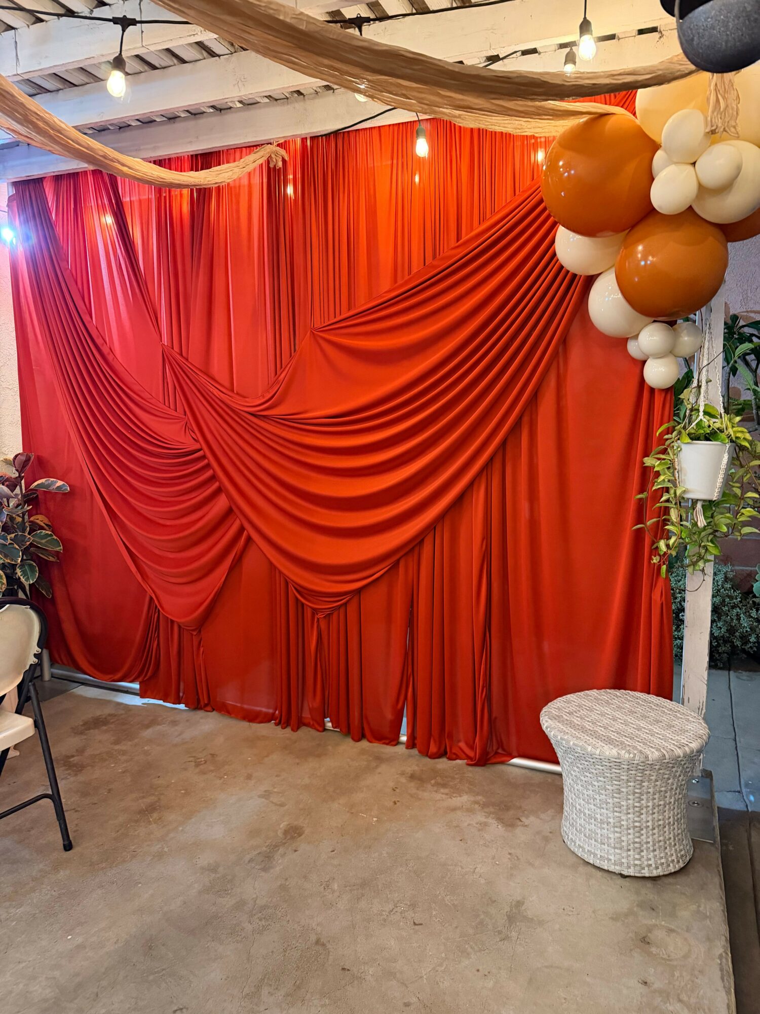 Red draped curtain with balloons and plants, decorated for celebration or event, with a wicker stool nearby.
