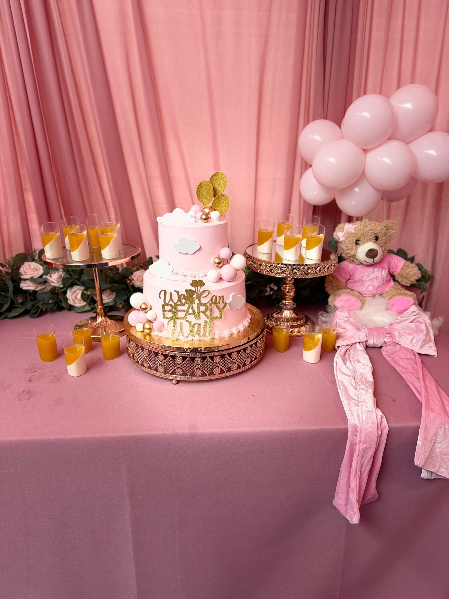 Pink birthday cake with bear decorations, surrounded by drinks, balloons, and a teddy bear on a pink table.
