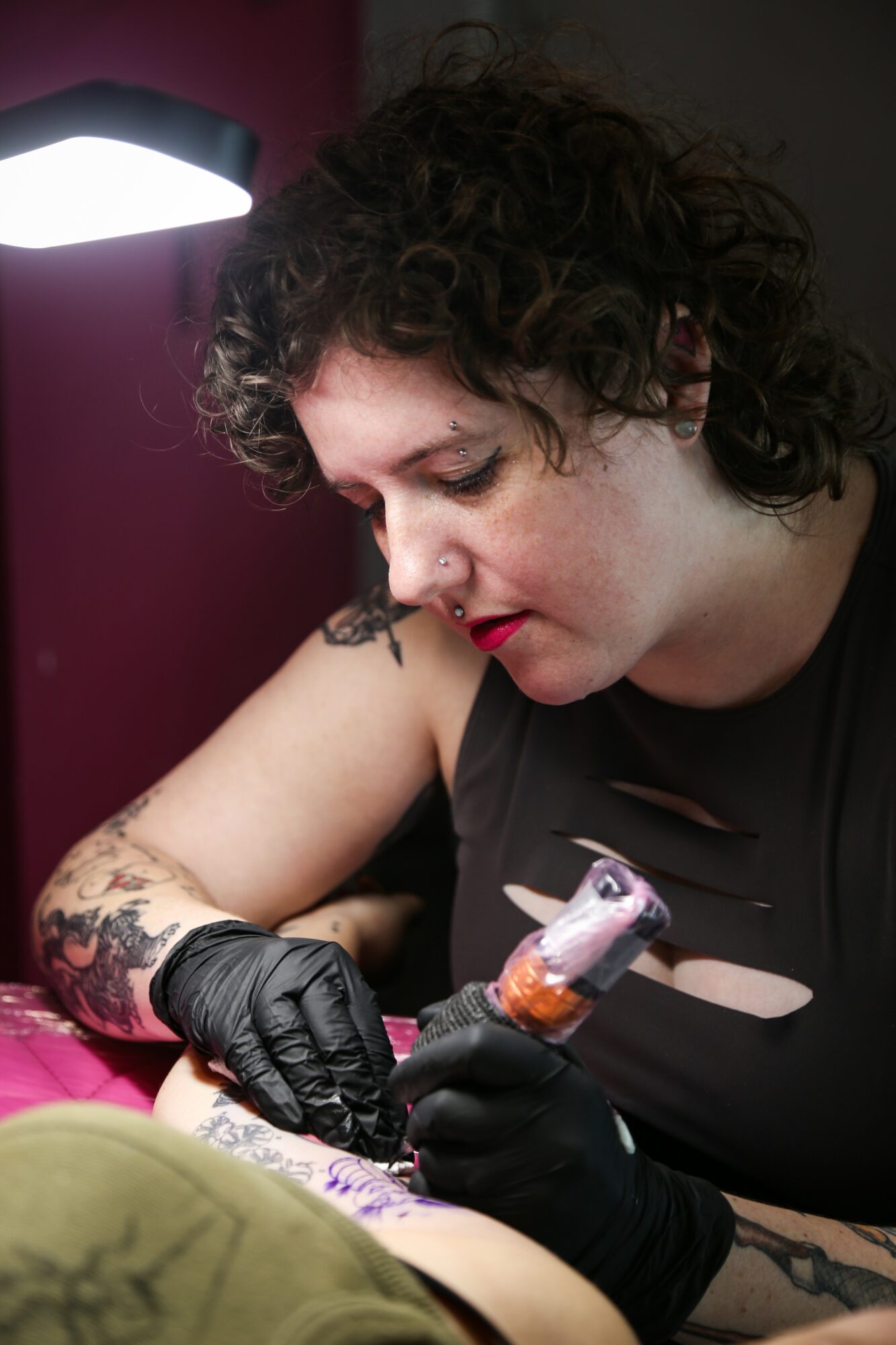 Woman with curly hair tattooing a person's arm, wearing black gloves and a black top, focused on her work.