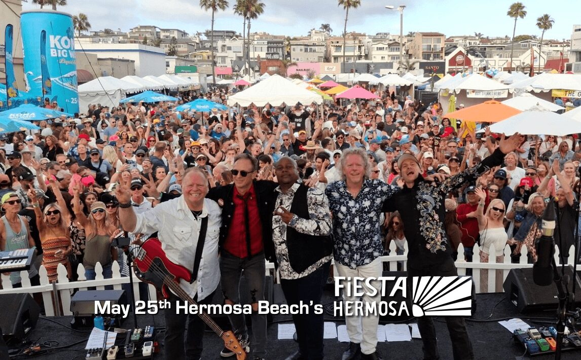Four men stand on stage in front of a large crowd at Hermosa Beach event, with tents and palm trees in background.