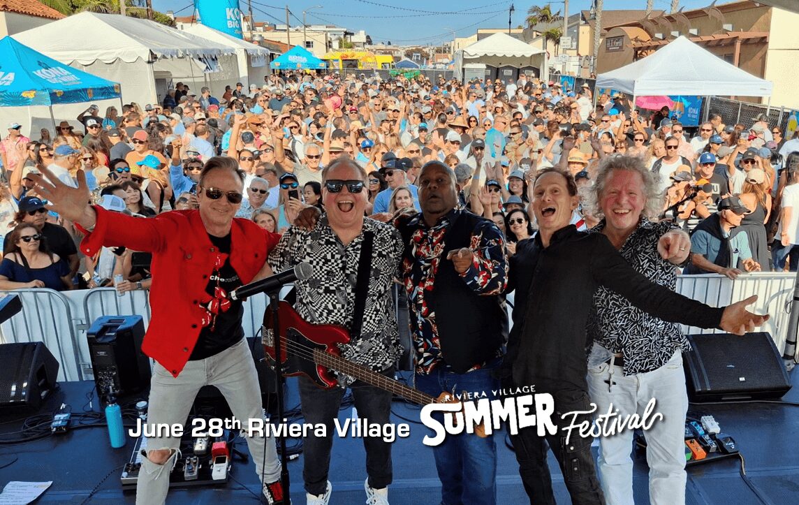 Four men on stage with arms outstretched, smiling, in front of a large crowd at an outdoor summer festival.