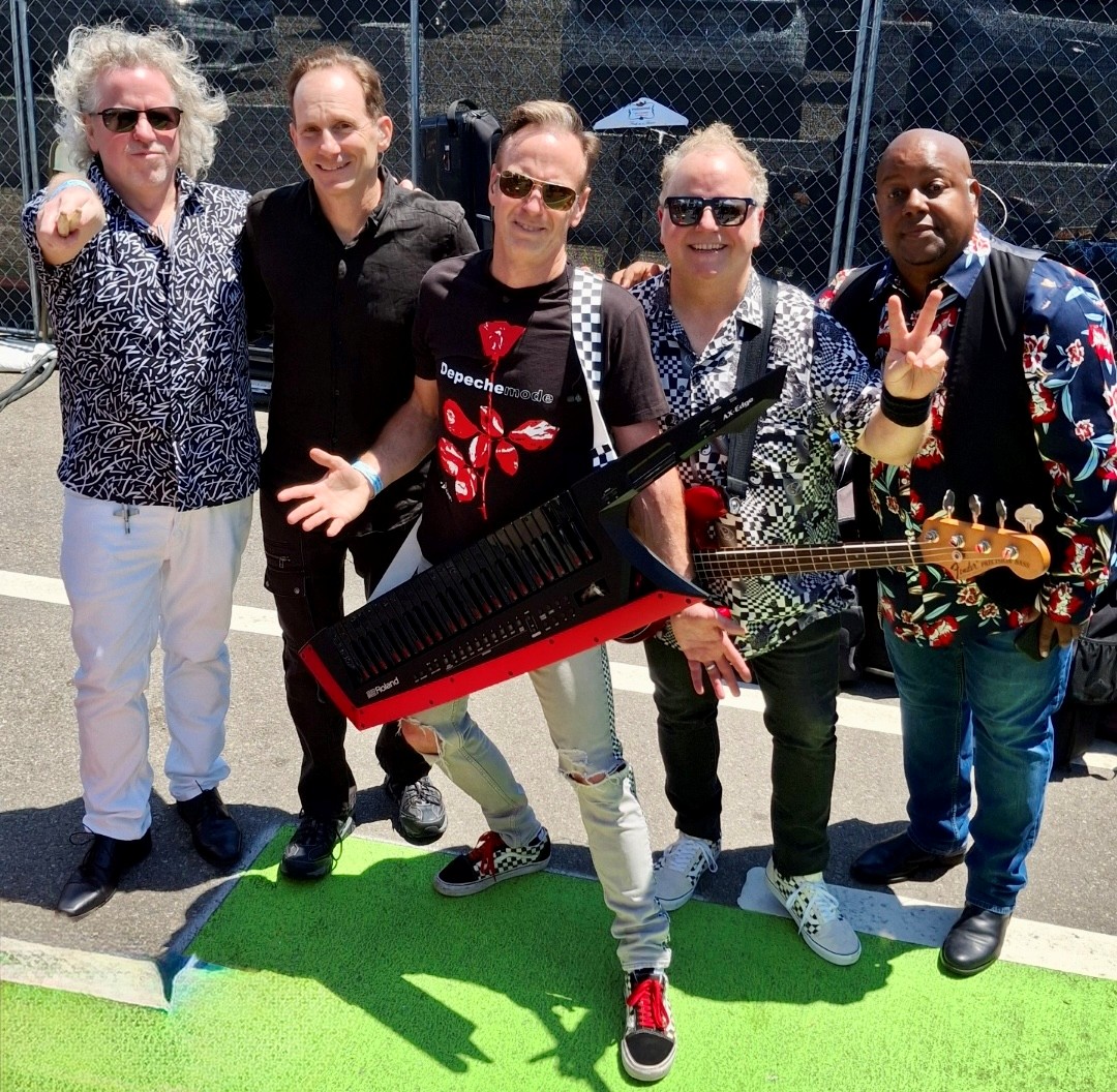 Group of five people standing outdoors, holding a large black and red guitar-shaped object, smiling, with a chain-link fence in background.