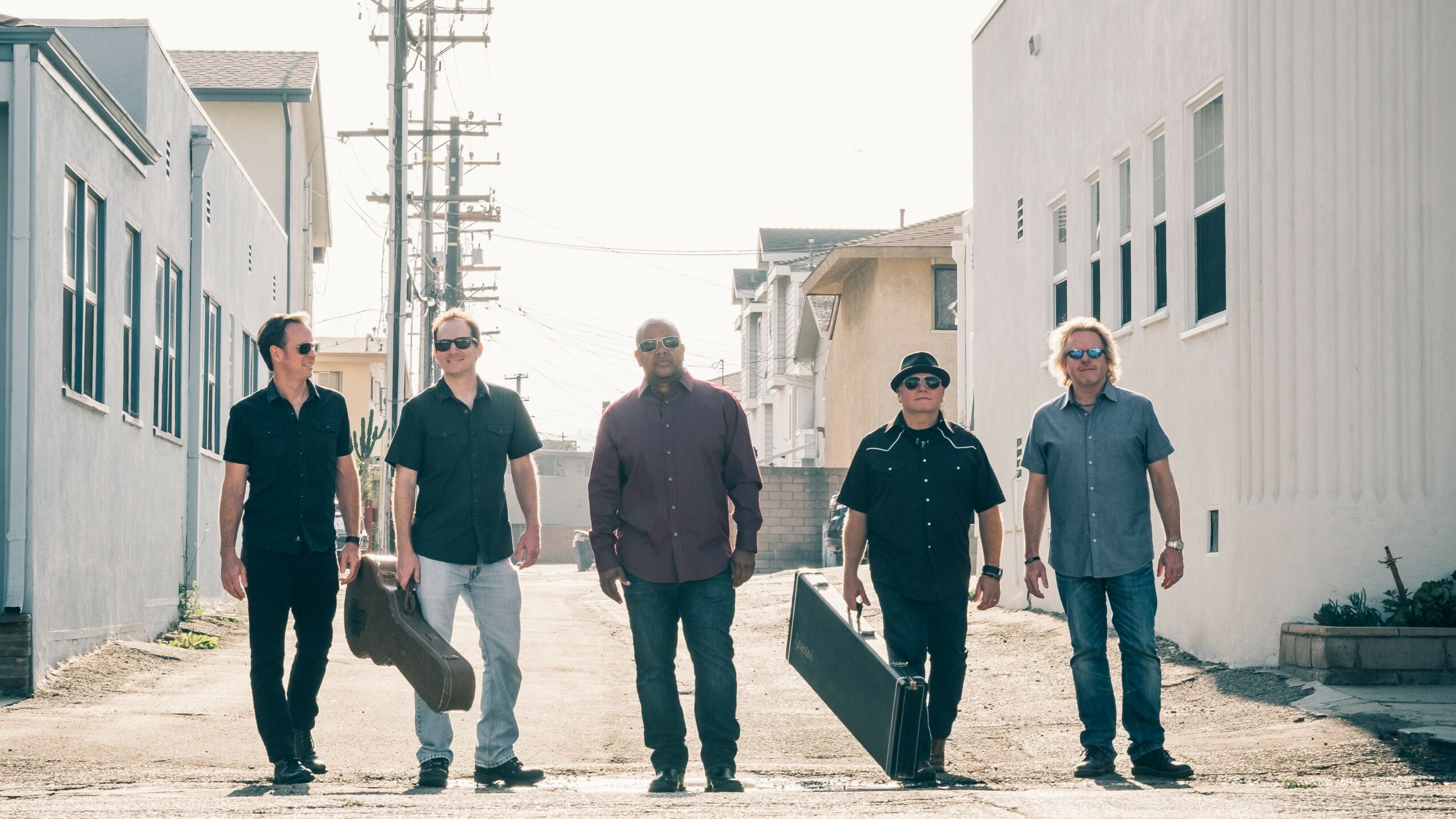 Five men walking down a street with buildings on both sides, some carrying cases, in casual and police attire.