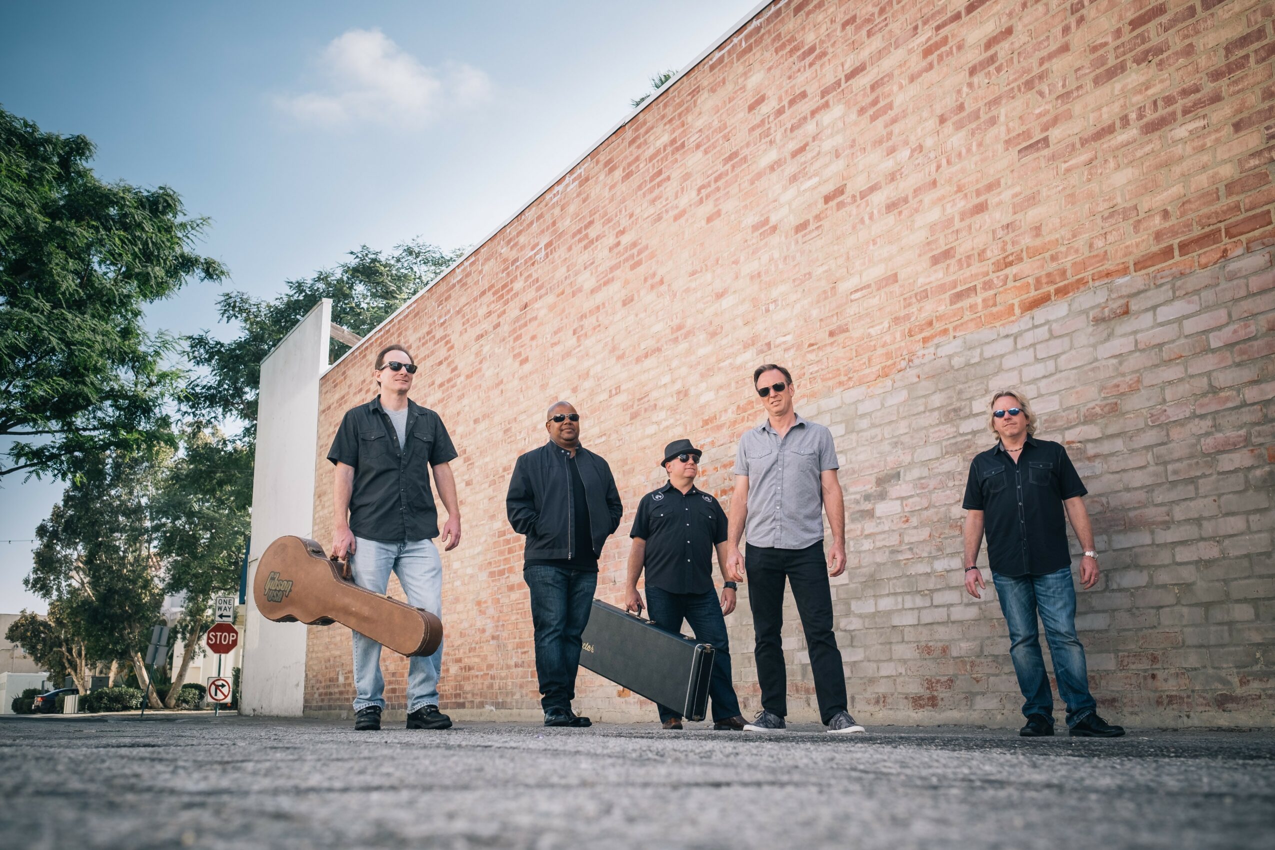 Five men walking on street with brick wall and trees in background, carrying musical instrument cases.