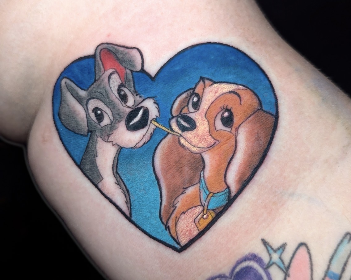 Tattoo of two animated dogs inside a heart shape, one gray and one brown, with a blue background.