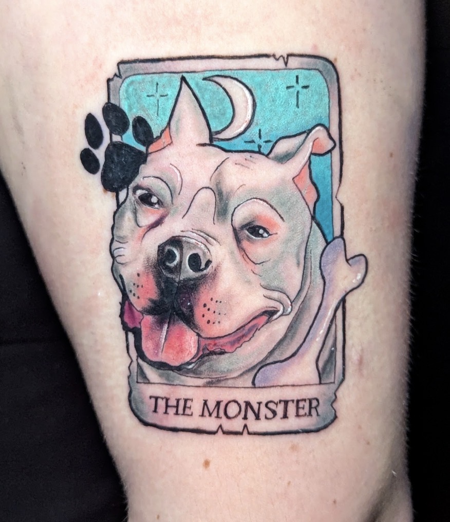 Dog tattoo with a moon, paw print, and banner reading 'The Monster'. Colorful design with a cartoon style.