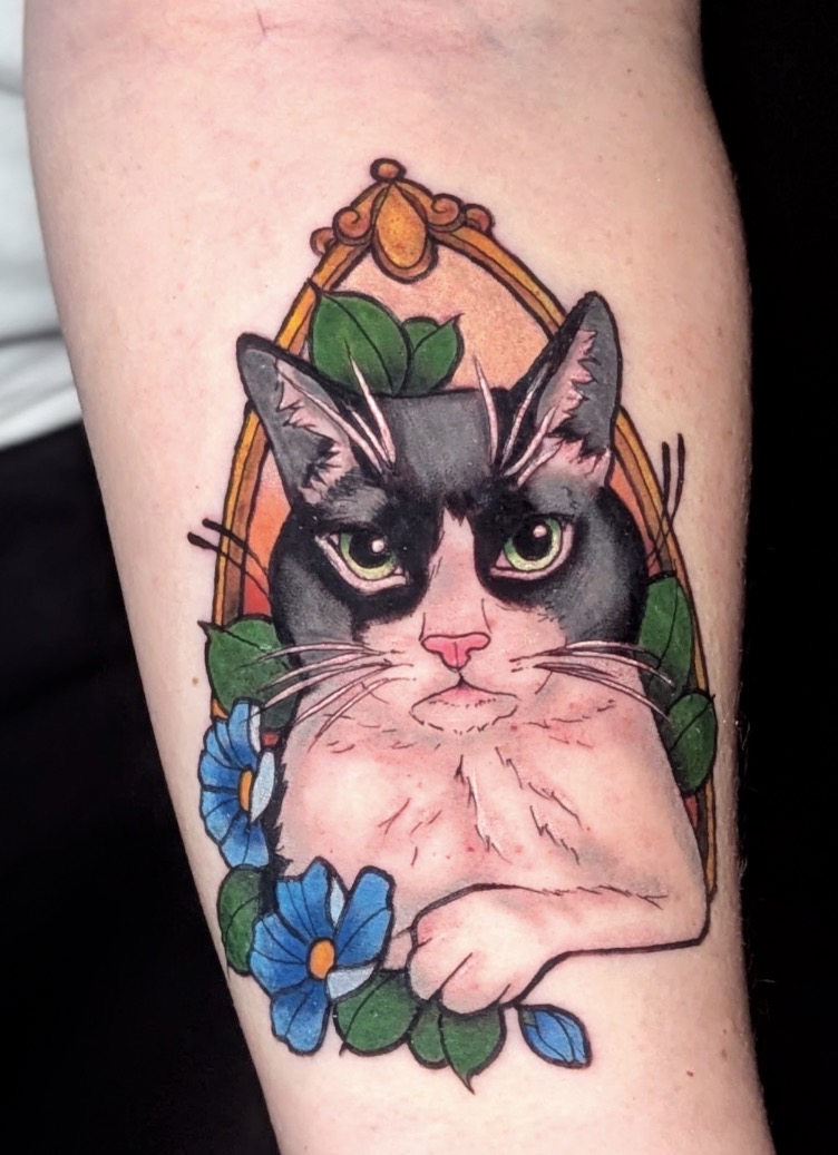 Tattoo of a cat's face with green eyes, surrounded by blue flowers and green leaves.