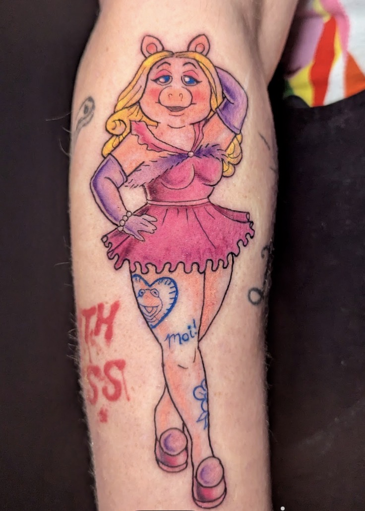 Tattoo of a cartoon pig girl with blonde hair, pink dress, and purple shoes, standing with one hand on her hip.