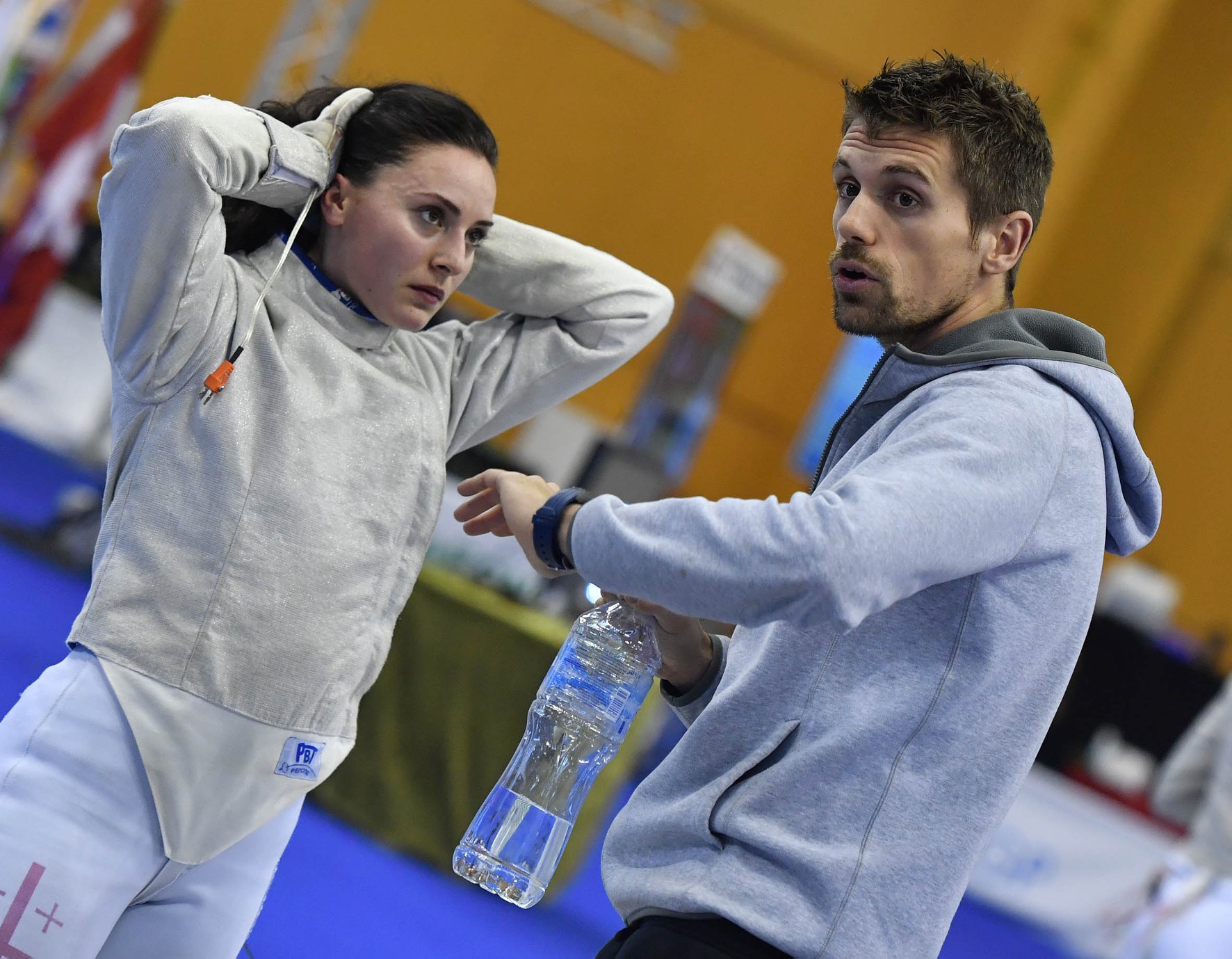 A woman in fencing attire listens to a man holding a water bottle, in an indoor sports setting.