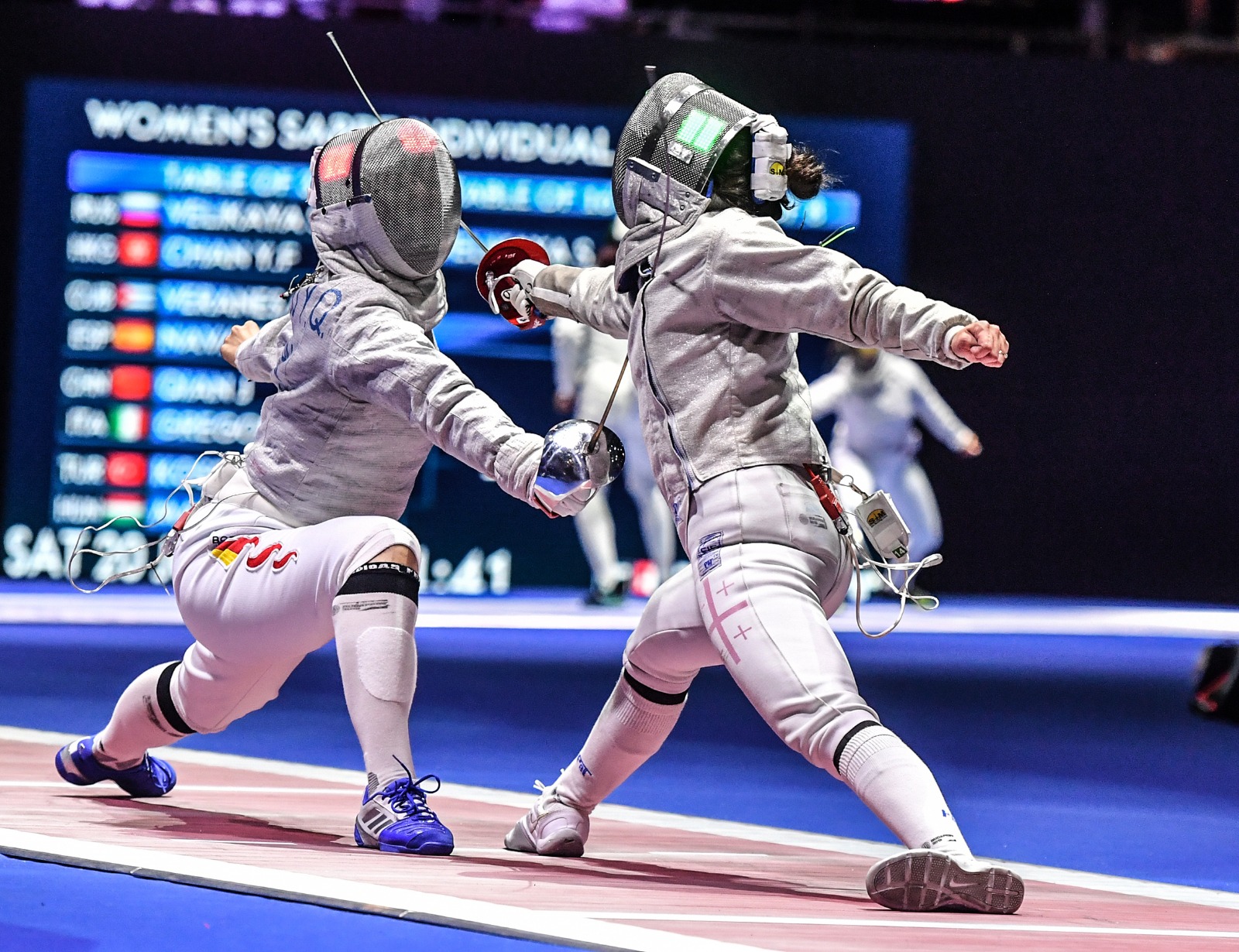 Two fencers in protective gear engage in a match on a blue fencing strip, with a scoreboard in the background.