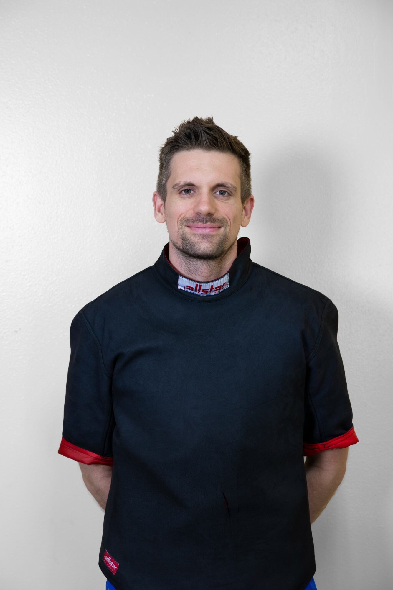 Man with short hair and beard wearing a black racing suit with red accents, standing against a plain wall.