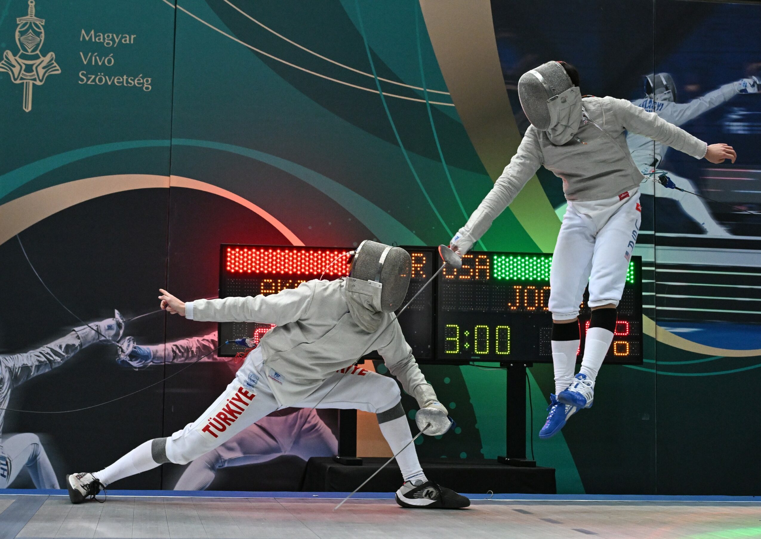 Two fencers in protective gear engage in a match, one lunging forward while the other blocks, on a fencing strip.