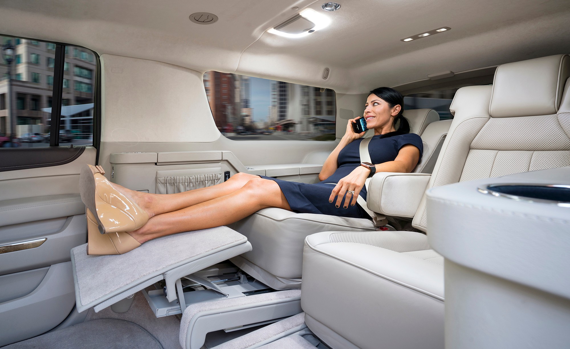 Woman sitting in luxury vehicle with legs stretched out, talking on phone, wearing dress and heels.