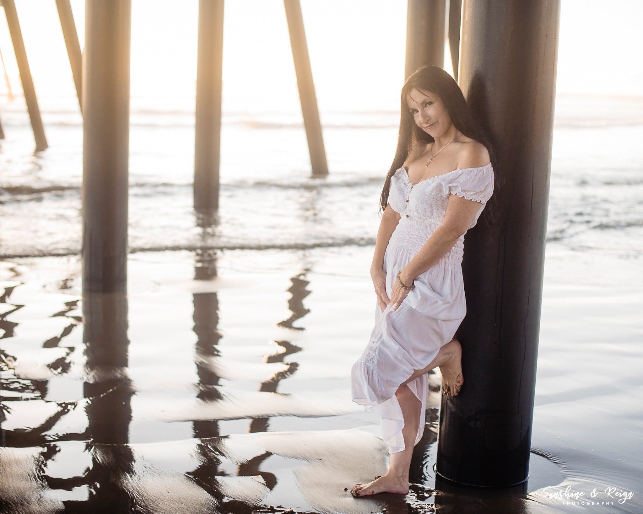 Young woman in white dress leaning against a pier post by the water, with sunlight and reflections on the surface.