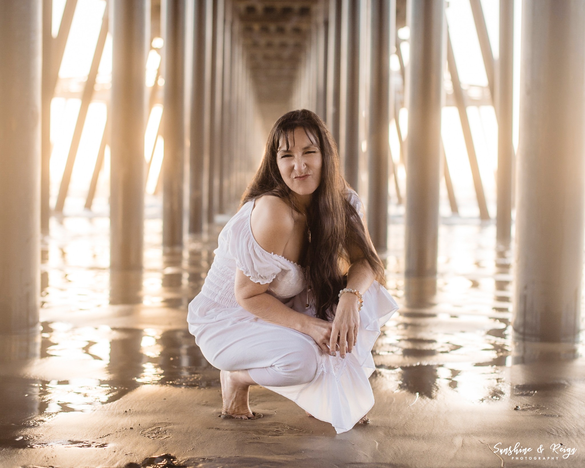 Woman crouching on wet surface under a bridge with metal pillars, sunlight shining through.