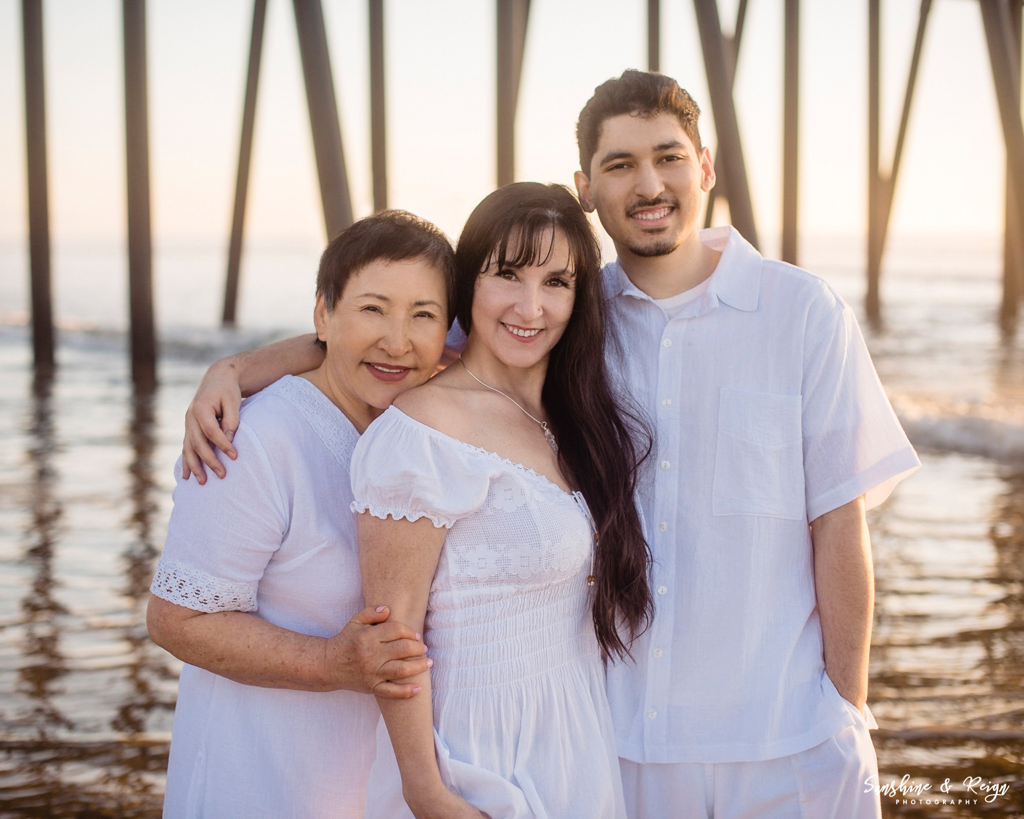 Three people smiling at the camera on a beach with a pier in the background during sunset.