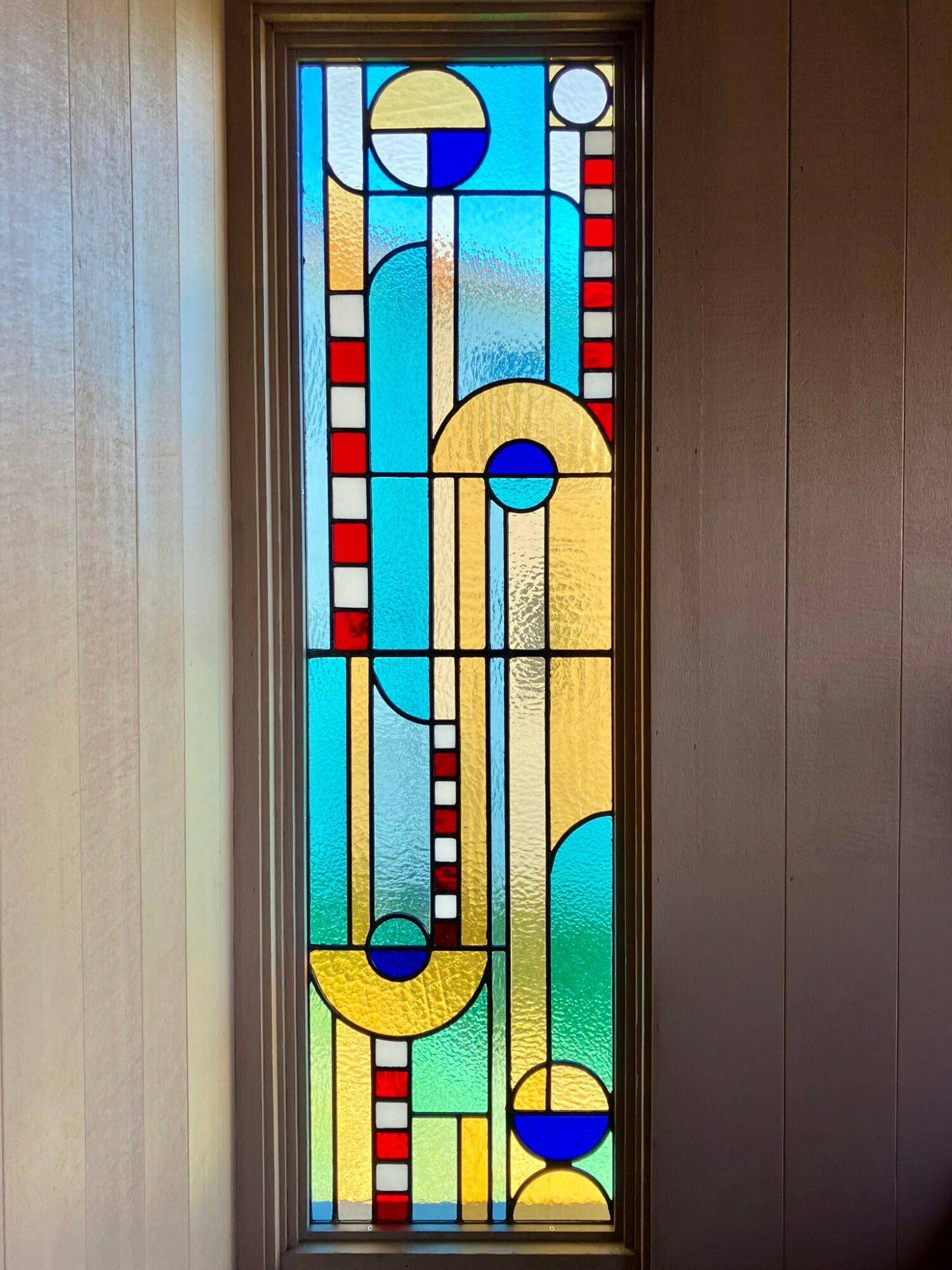 Vertical stained glass window with geometric shapes and colors including blue, yellow, red, and clear glass sections.