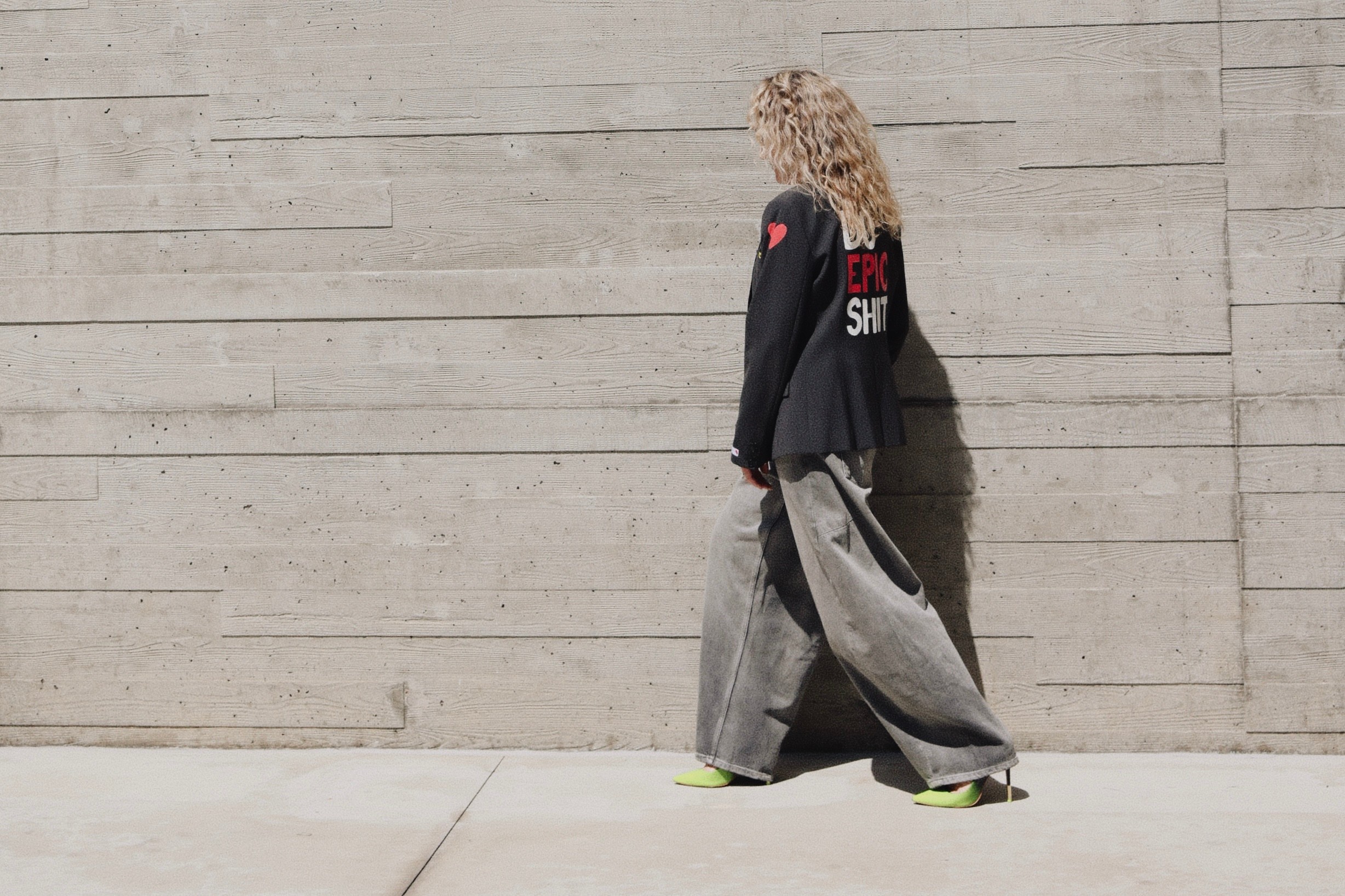 Person with curly hair standing against a concrete wall, wearing a dark jacket and wide pants.