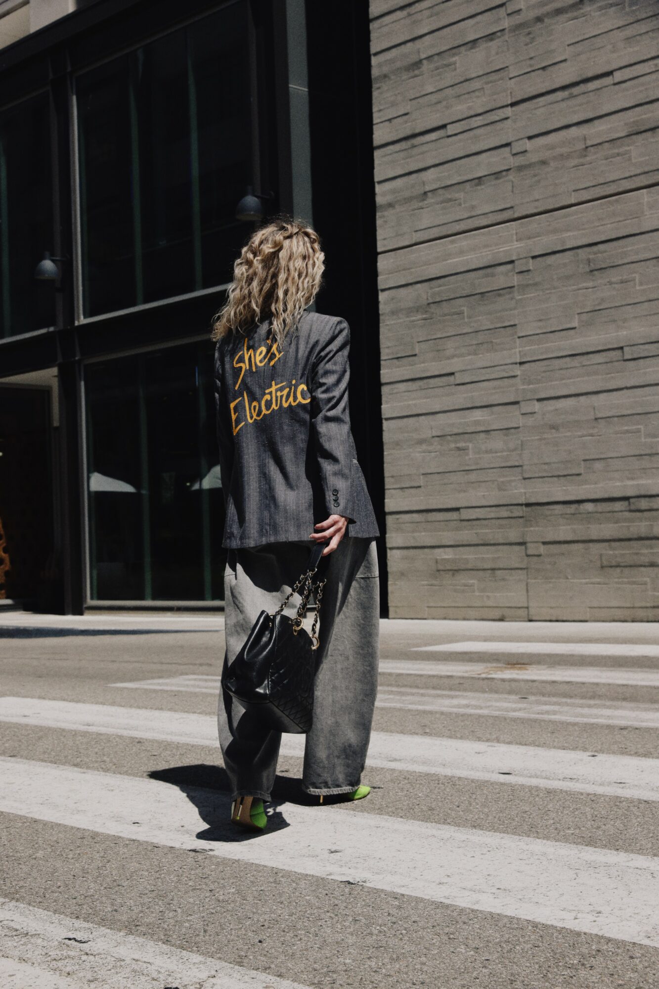 Person with curly hair walking across a street, carrying a bag, wearing a jacket and wide pants.