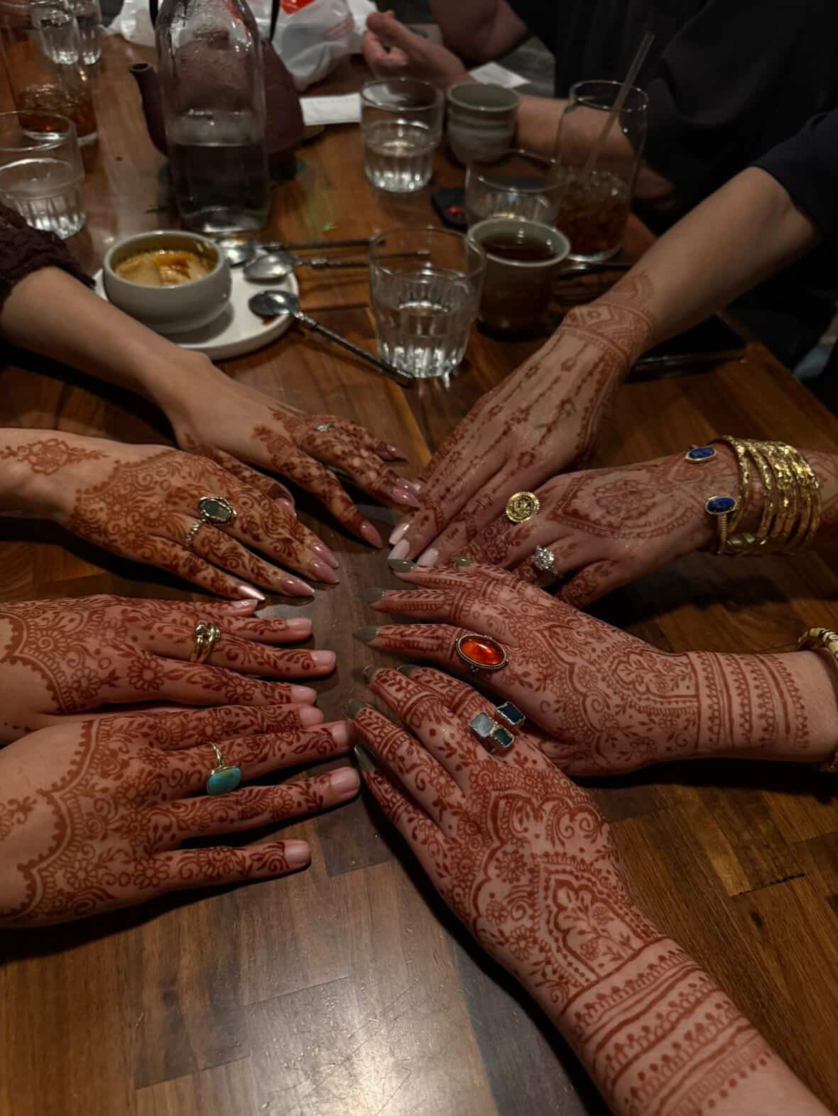 Multiple hands with henna designs and jewelry reach towards the center on a wooden table with drinks and bowls.
