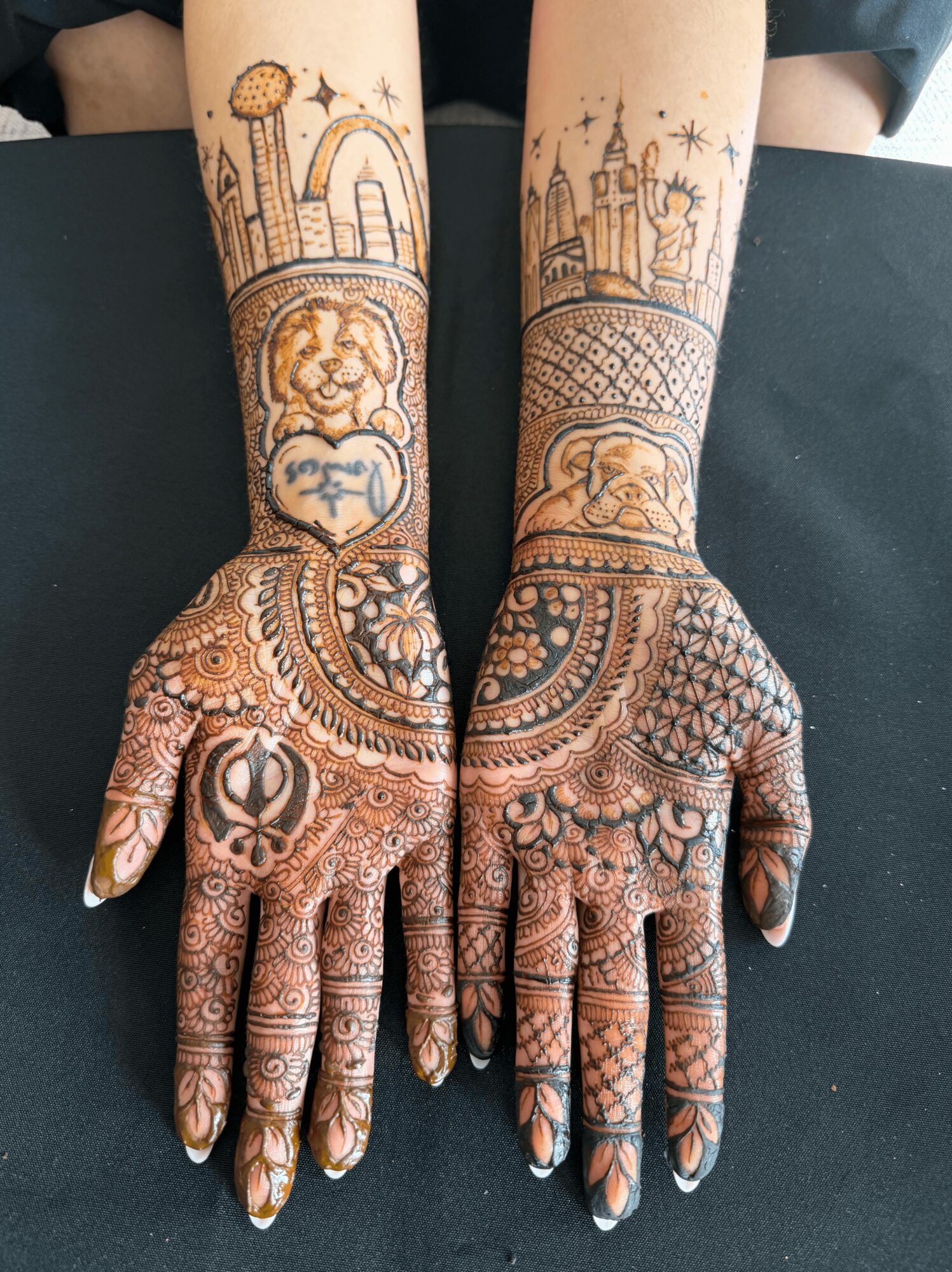 Two hands with intricate henna designs, extending to forearms with cityscape and architectural patterns, on a black surface.