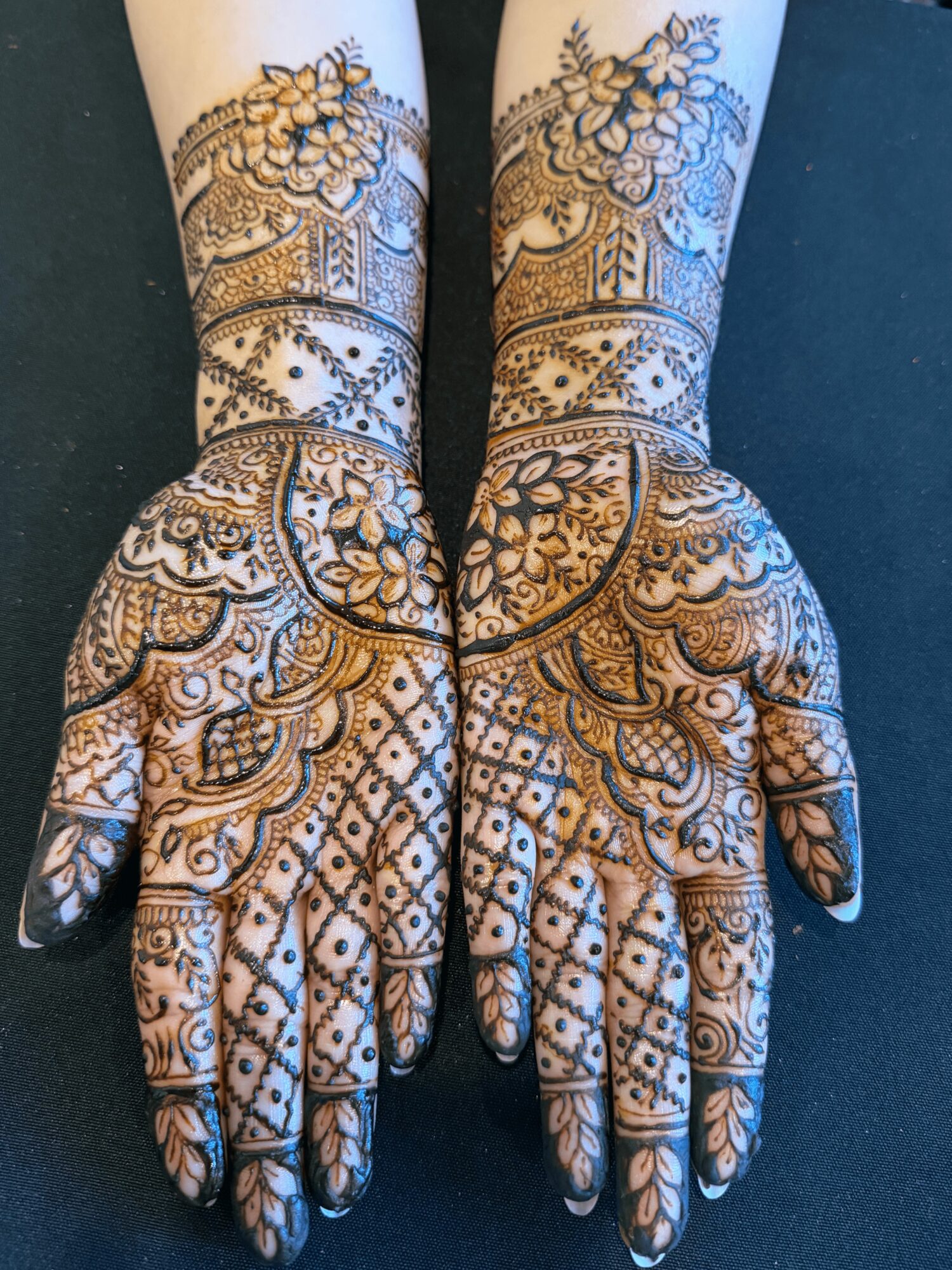 Two hands with intricate henna designs, extending to arms, featuring floral and geometric patterns on dark skin.