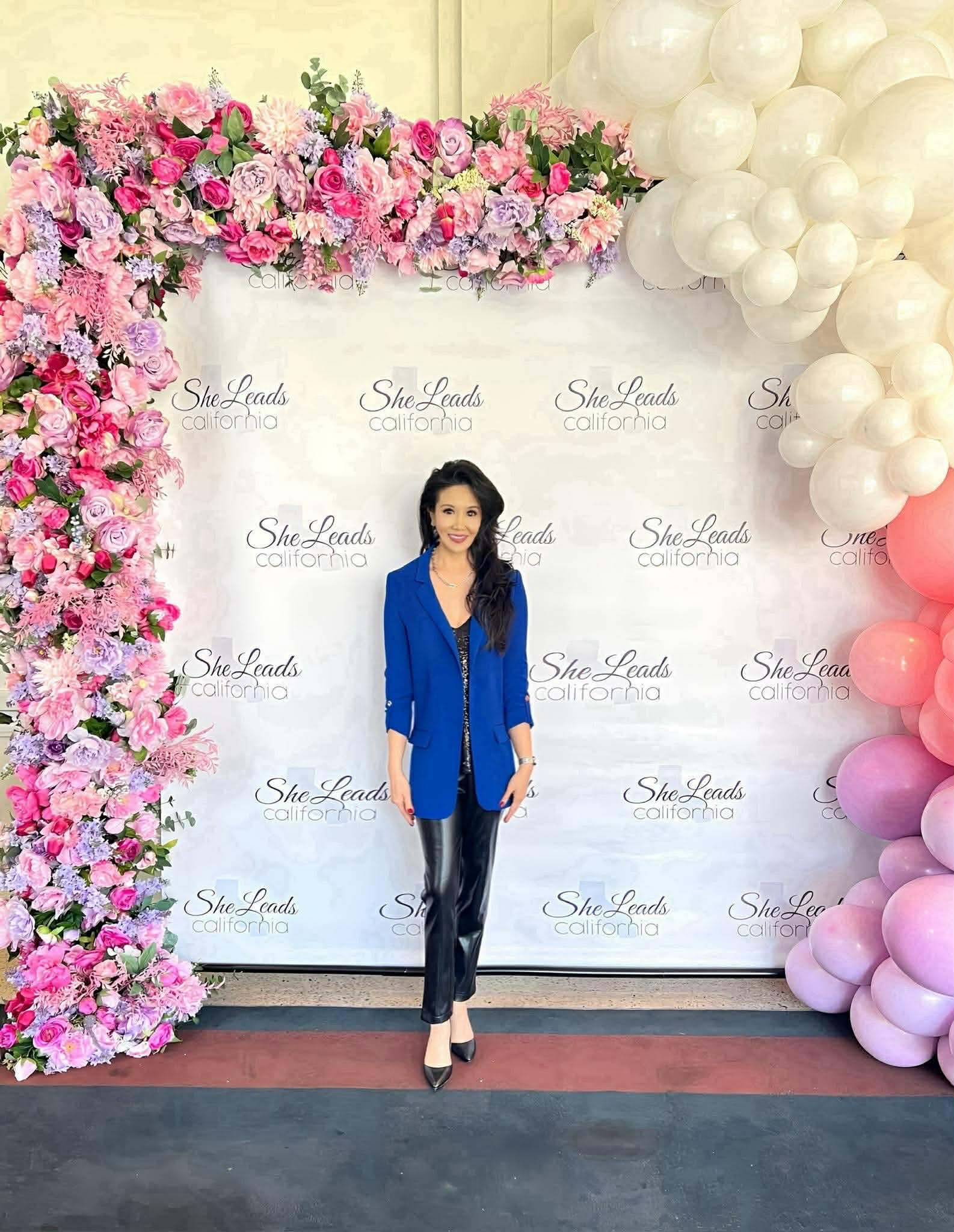 Woman in blue blazer and black pants standing in front of floral and balloon backdrop.