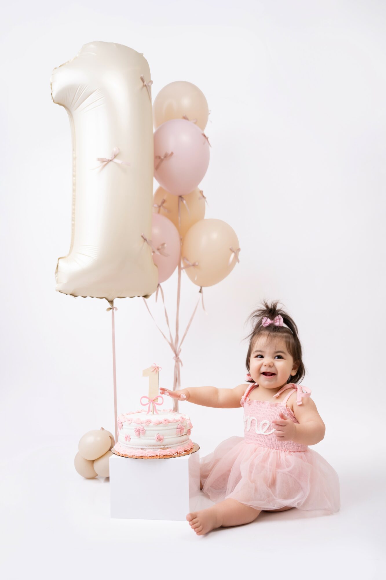 Child in pink dress sitting next to a birthday cake with balloons and a large number one balloon.