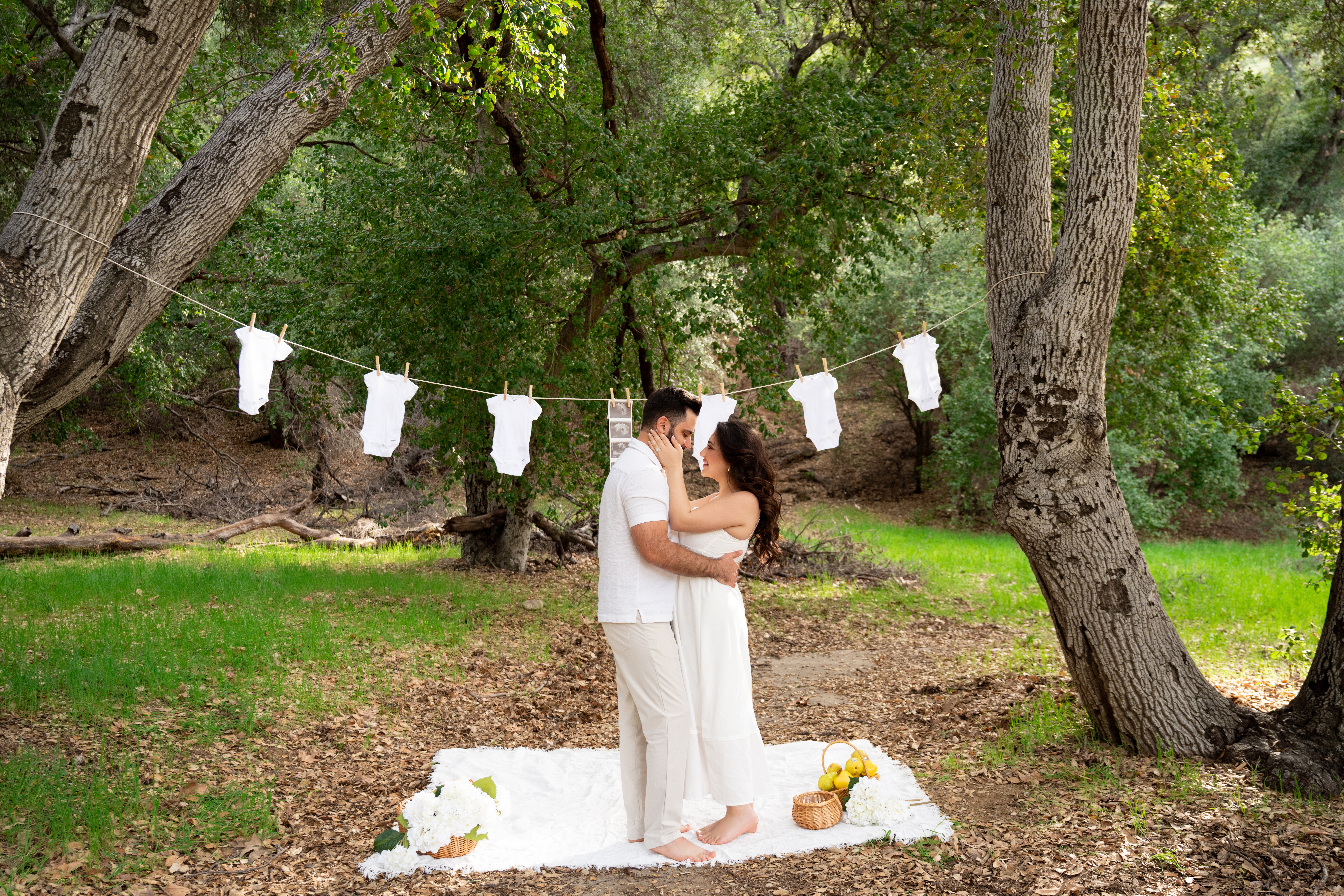 Couple embracing outdoors under trees with white paper lanterns hanging above them, on a grassy area with a white cloth and flowers.