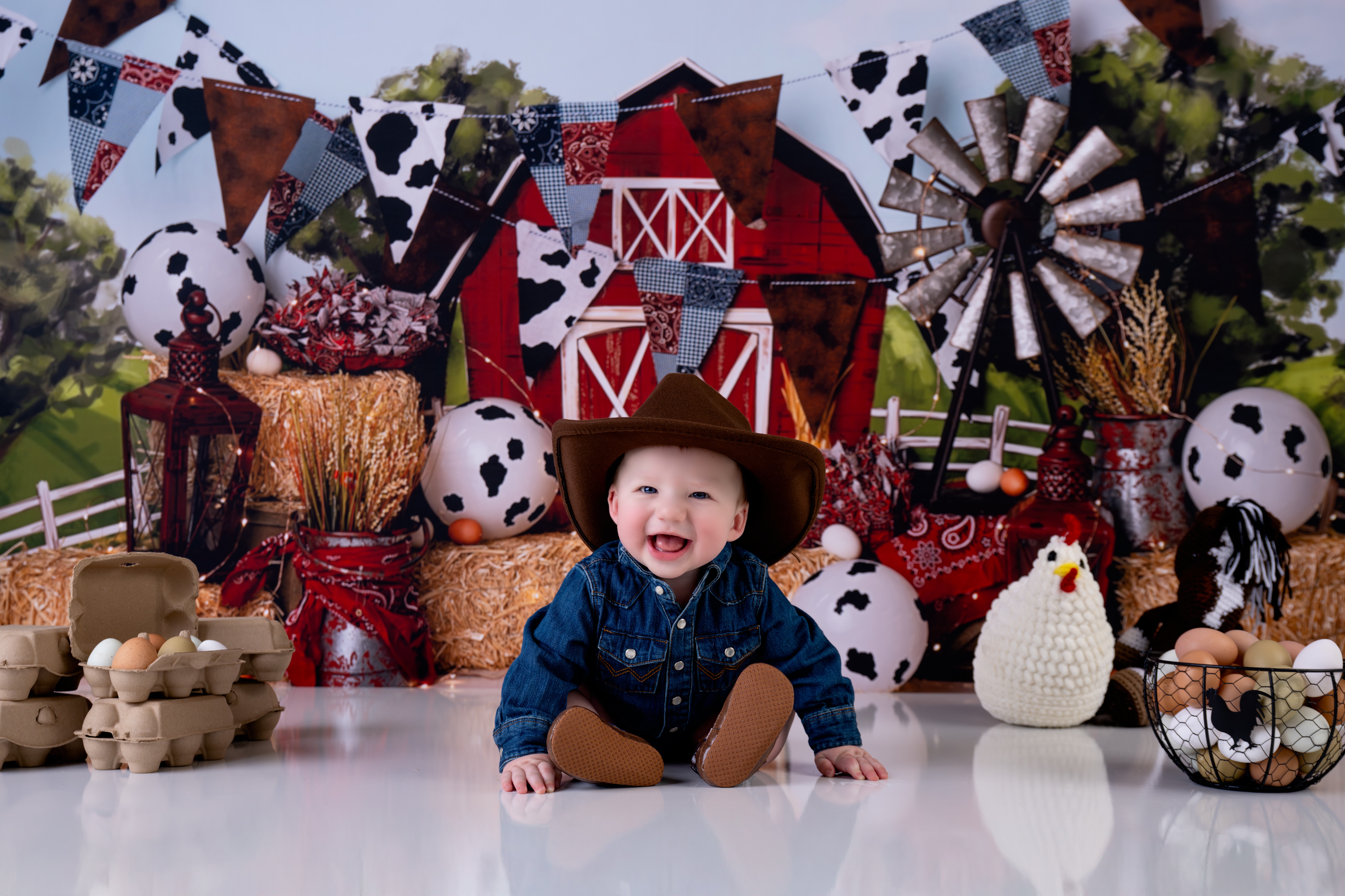 Child dressed as a cowboy, smiling, sitting on a decorated farm-themed background with hay bales and farm props.