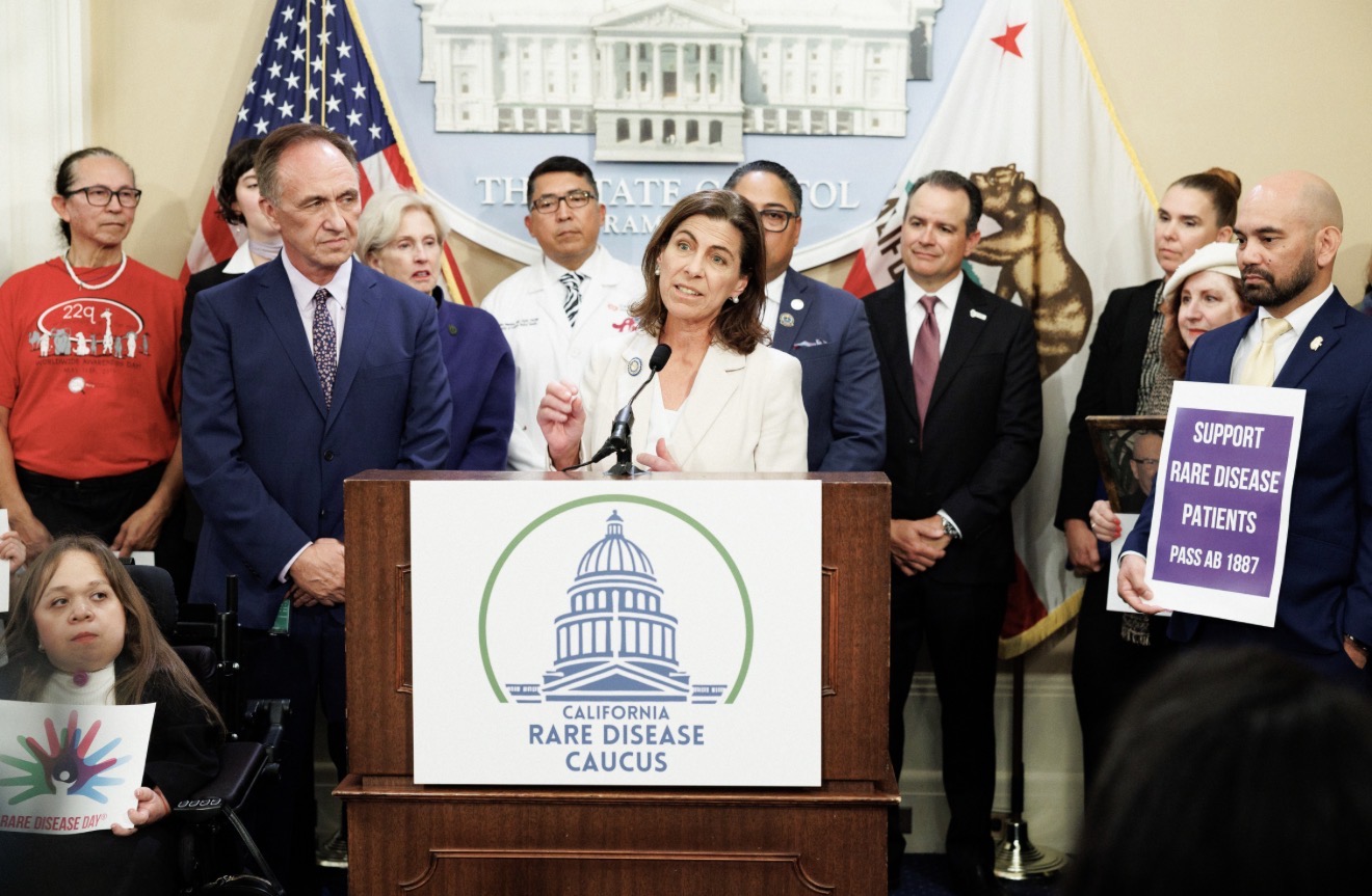 Group of people standing behind a podium with a sign reading 'California Rare Disease Caucus,' in a government room.