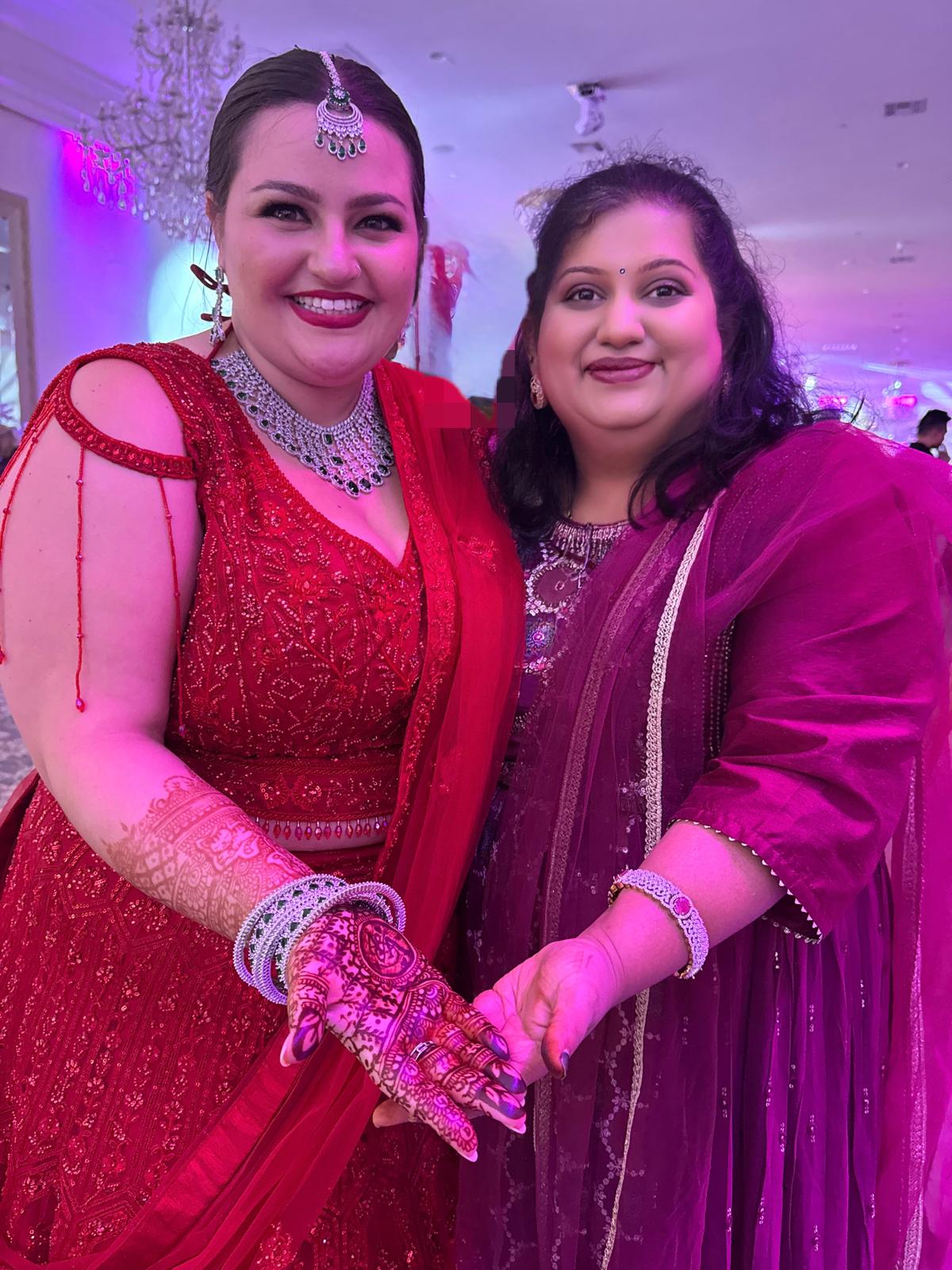 Two women in traditional attire smiling, holding hands, at a festive event with colorful lighting.